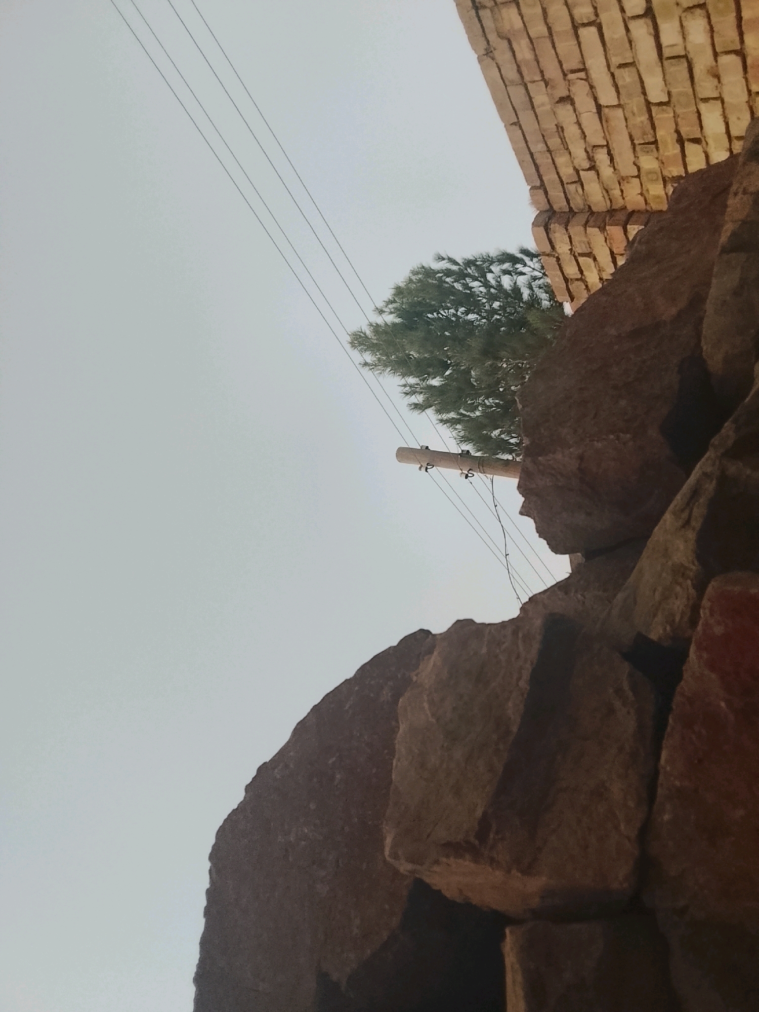 An outdoor, low-angle shot of a cluster of large rocks in Paldorak, Tajikistan. The rocks are a mixture of dark brown and red hues and are piled high, obscuring most of the background. In the upper right corner, a wall constructed of tan bricks is visible, also appearing aged and weathered. Above the rocks and brick wall, the sky is a pale, hazy gray. A single, thin evergreen tree with dark green foliage is positioned behind the rocks, its branches reaching towards the sky. Four thin, dark power lines stretch diagonally across the frame from the upper left to the middle right. A wooden utility pole, weathered and gray, is located behind the rocks and tree, with insulators attached to it.
