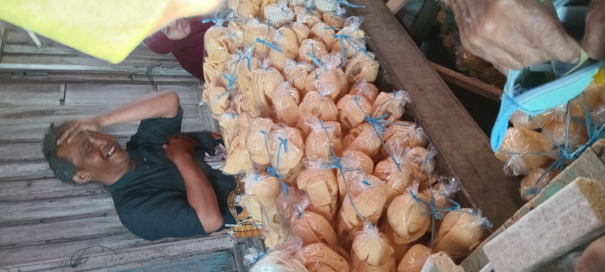 An outdoor market stall in Sindangwangi, Indonesia displays a large pile of snack crackers. The crackers are individually wrapped in clear plastic bags, tied with blue string, and are a light golden-yellow color. In the foreground, a person's hands are visible, reaching towards a light blue bag. To the left, a block of tempeh, a fermented soybean product, wrapped in white paper, is also on display. Behind the crackers, a man with grey hair is laughing heartily, with his hand on his forehead. He is wearing a dark blue shirt and a patterned sarong. Further in the background, a woman wearing a maroon hijab is visible, looking towards the right. The stall is constructed of weathered wood, suggesting an established market setting.