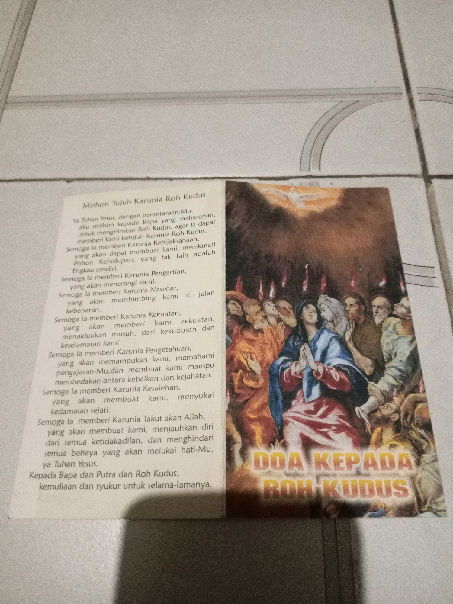 The image shows a printed prayer with a religious painting. The prayer, titled "Mohon Tujuh Karunia Roh Kudus," is written in Indonesian. The painting depicts a scene with multiple figures, many of whom appear to be looking upwards. A dove, representing the Holy Spirit, is shown descending from the heavens. The text beneath the painting reads "DOA KEPADA ROH KUDUS," which translates to "Prayer to the Holy Spirit." The photograph was taken indoors, with the prayer and painting lying flat on a tiled surface. The tiles are white with grey grout lines. The lighting suggests an indoor setting, possibly a home or a place of worship. The overall scene is one of religious devotion and imagery. There is no indication of the specific location in the city of Malang, Indonesia.