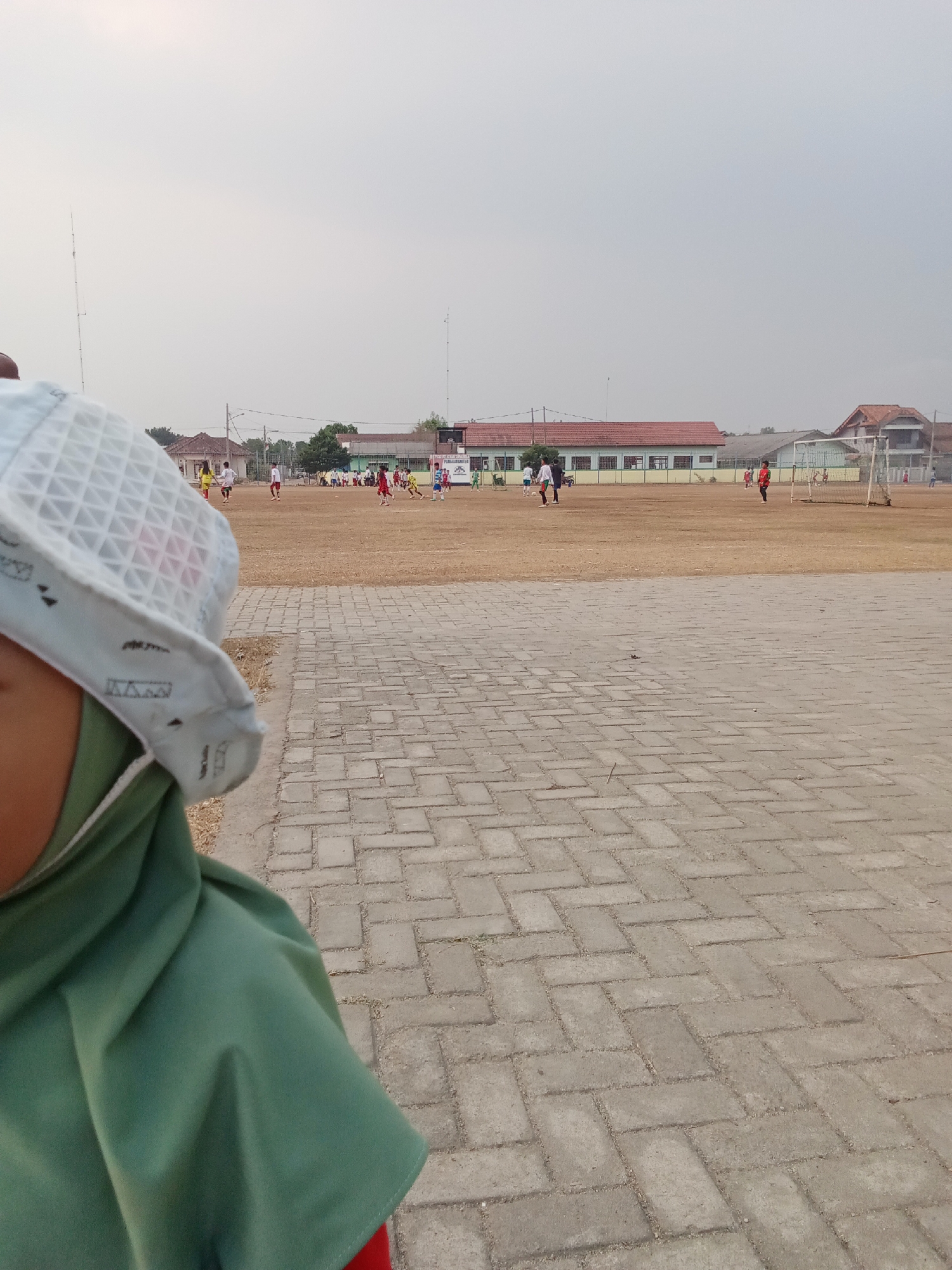 The image depicts a daytime scene at what appears to be a sports field, possibly in Toto Katon, Indonesia. In the foreground, a person wearing a green hijab and a white patterned hat is partially visible, looking towards the right. The background reveals a dusty, open field with several figures engaged in a soccer game. Buildings with red roofs line the far edge of the field, and some trees are scattered around the area. A soccer goal is visible on the right side of the frame. The ground in the foreground is paved with rectangular grey bricks arranged in a herringbone pattern. The sky is overcast and appears hazy.