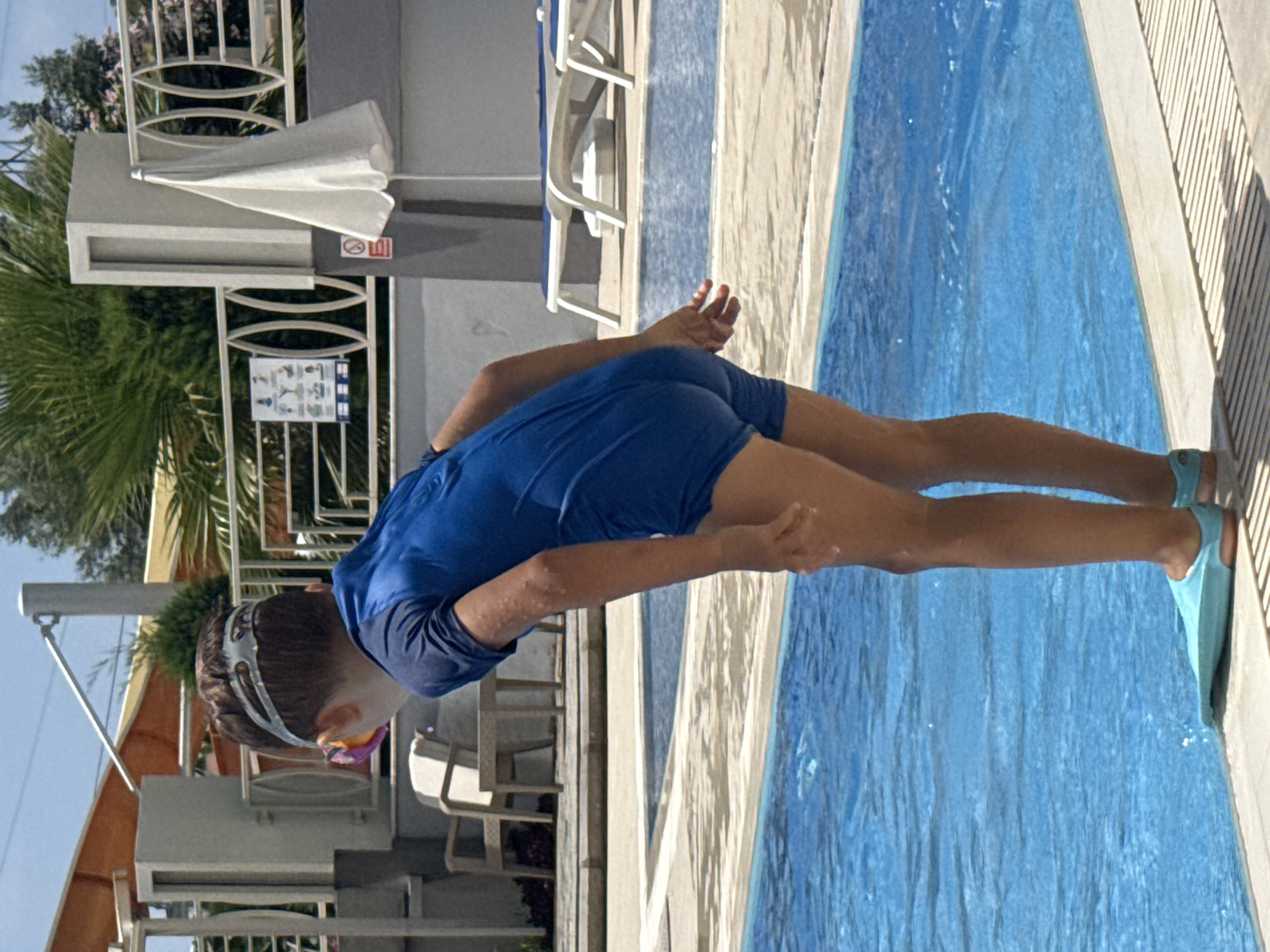 The image shows a young person wearing a blue rash guard, goggles, and flippers, leaning over the edge of a swimming pool. The person is positioned to enter the water, with their body bent at the waist and arms extended towards the pool. The pool water is a vibrant blue, and sunlight reflects off its surface.
The setting appears to be an outdoor swimming pool area, likely at a resort or hotel, given the presence of lounge chairs and a fence in the background. The architecture suggests a Mediterranean or similar warm climate, consistent with the provided location context of Alanya, Turkey. Lush green palm trees and other foliage are visible, indicating it's likely daytime and the weather is pleasant. The person's posture and gear suggest they are about to jump or dive into the pool for recreational swimming or perhaps a diving activity. Their gaze is directed downwards towards the water, and their body is tensed in anticipation.
On a panel in the background, there is a warning sign with red and white symbols, and another informational poster with illustrations of swimming pool safety rules. No discernible text is clearly legible beyond these visual cues. The overall mood is one of anticipation and readiness for aquatic fun.
