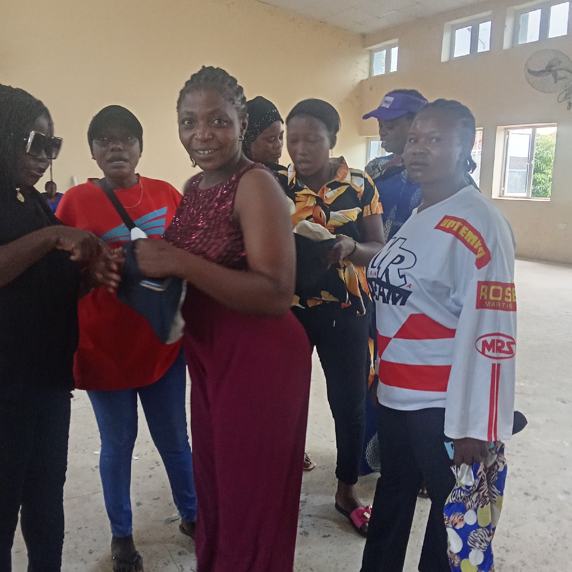 A group of women stand inside a building. The location is likely in Akwanga, Nigeria, based on contextual information not provided in the image. The building has light-colored walls, several windows, and a ceiling fan. The women are wearing a variety of clothing: dresses, pants, shirts, and caps. One woman wears sunglasses and a black outfit. Another wears a red shirt and blue jeans. One wears a maroon dress. A fourth wears a white long-sleeved shirt with red and black markings. Some women are holding bags. They appear to be in conversation.