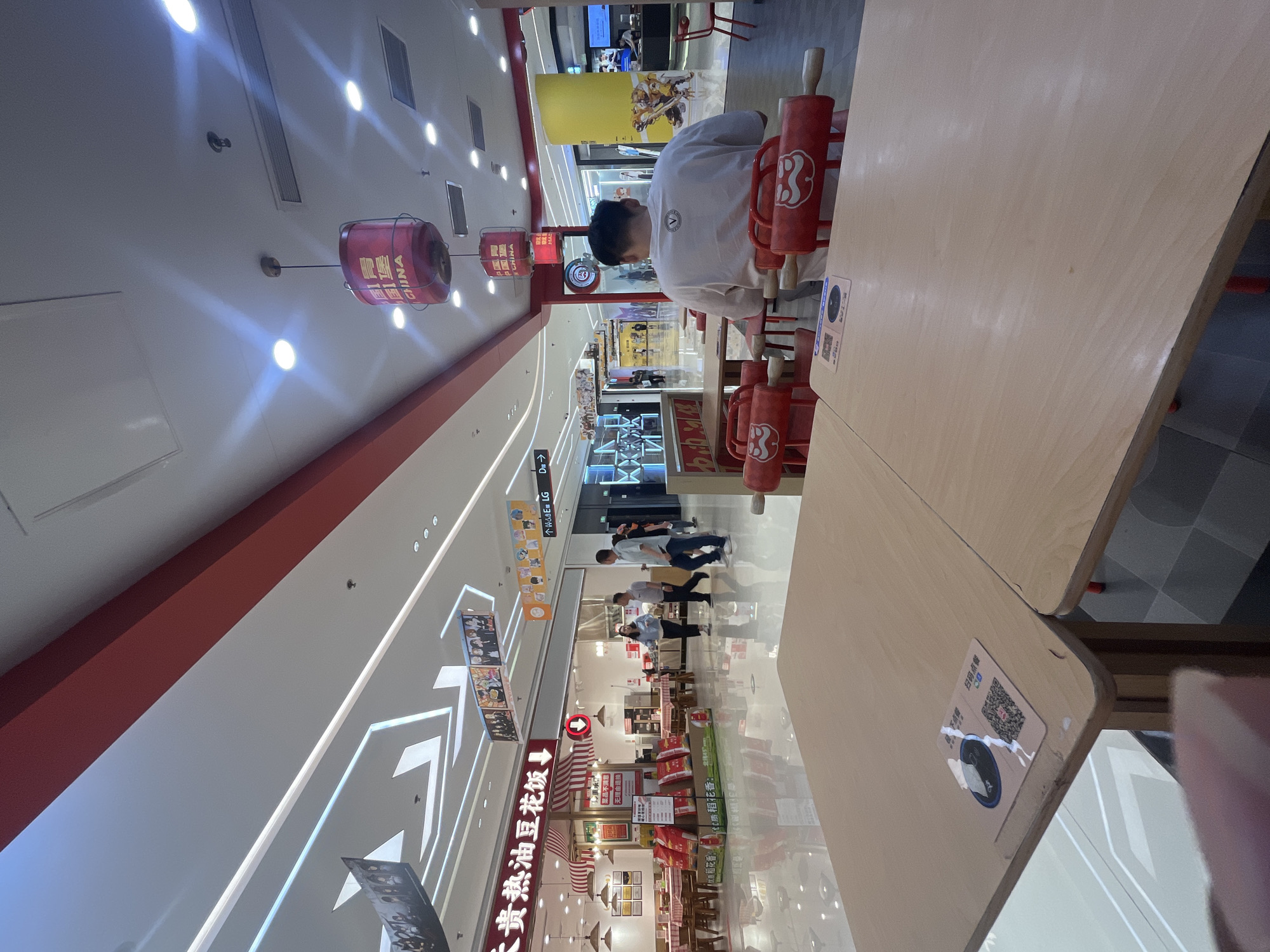 This is a medium shot, taken from a slightly low angle, of a food court or restaurant interior in a shopping mall. The scene is well-lit with overhead fluorescent lighting and decorative lamps. In the foreground, two light-colored wooden tables are visible, with one in focus and the other slightly blurred. On the focused table, a QR code sticker is placed towards the left. The table is set with red chairs that have distinctive rolling pin-shaped backrests with a cartoonish mustache design.
In the middle ground, a man in a white t-shirt with a logo on the back is seated at one of the tables, facing away from the camera. Beyond him, a restaurant with a red and white striped awning is visible. The restaurant has tables set with red and white checkered cloths and bags of merchandise on display. Several people are walking in the background, and there are various shopfronts and displays, including a yellow cylindrical pillar and posters with animated characters. Decorative red lanterns with Chinese characters are suspended from the ceiling. The floor is highly reflective, mirroring the ceiling lights and the surrounding environment. The overall atmosphere suggests a busy public space, likely a shopping mall in the city of Yuzhong District, China, given the visible Chinese text and the architectural style.