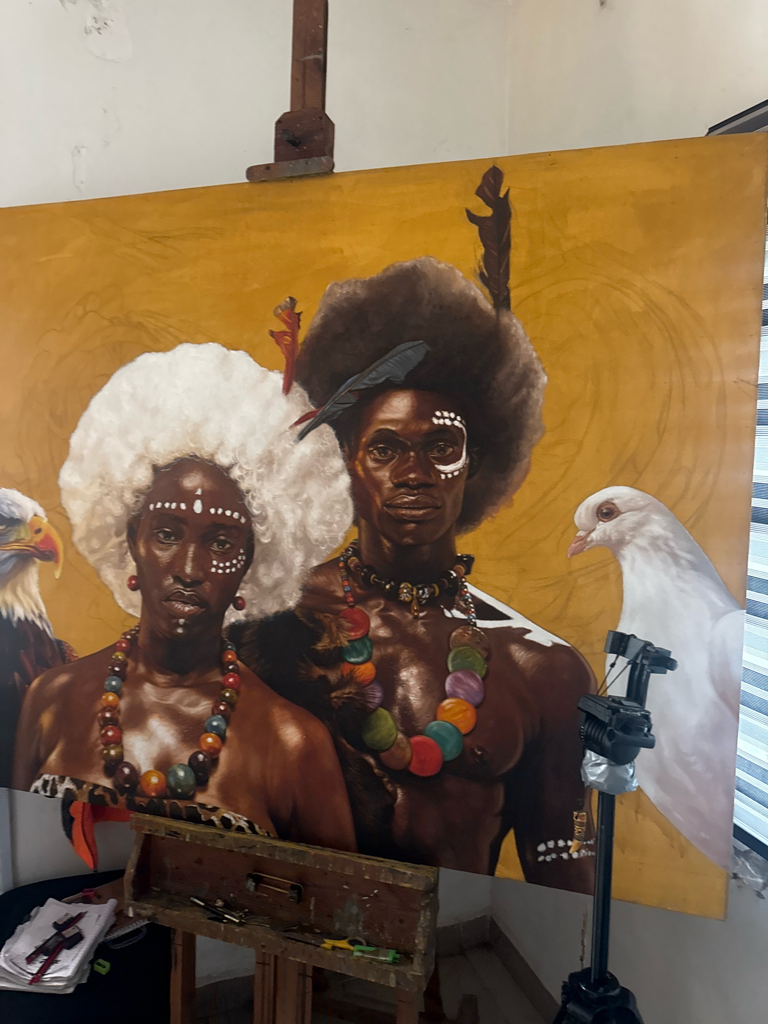 This is a photograph of a large oil painting depicting two figures, likely a man and a woman, adorned with traditional markings and jewelry. The figures have dark skin and elaborate hairstyles. The woman wears a voluminous white afro, while the man has a large, dark afro. Both wear colorful beaded necklaces. The man also has facial markings, and a small bird is perched on his head.
To the left of the figures is a detailed painting of an eagle, and to the right is a white dove. The background of the painting is a warm, golden-yellow hue with subtle circular patterns. The painting is displayed on an easel in what appears to be an artist's studio. In the foreground, a wooden artist's toolbox is visible, containing brushes and other art supplies. A tripod with a camera mounted on it is positioned in front of the right side of the painting, suggesting the artwork is being documented or displayed. The overall setting is indoors, with striped blinds visible in the background to the right. The presence of an easel, art supplies, and a large unfinished canvas indicates an active artistic workspace. The image itself does not provide direct information about its location in Ado, Nigeria, beyond the context of the artwork's subject matter.