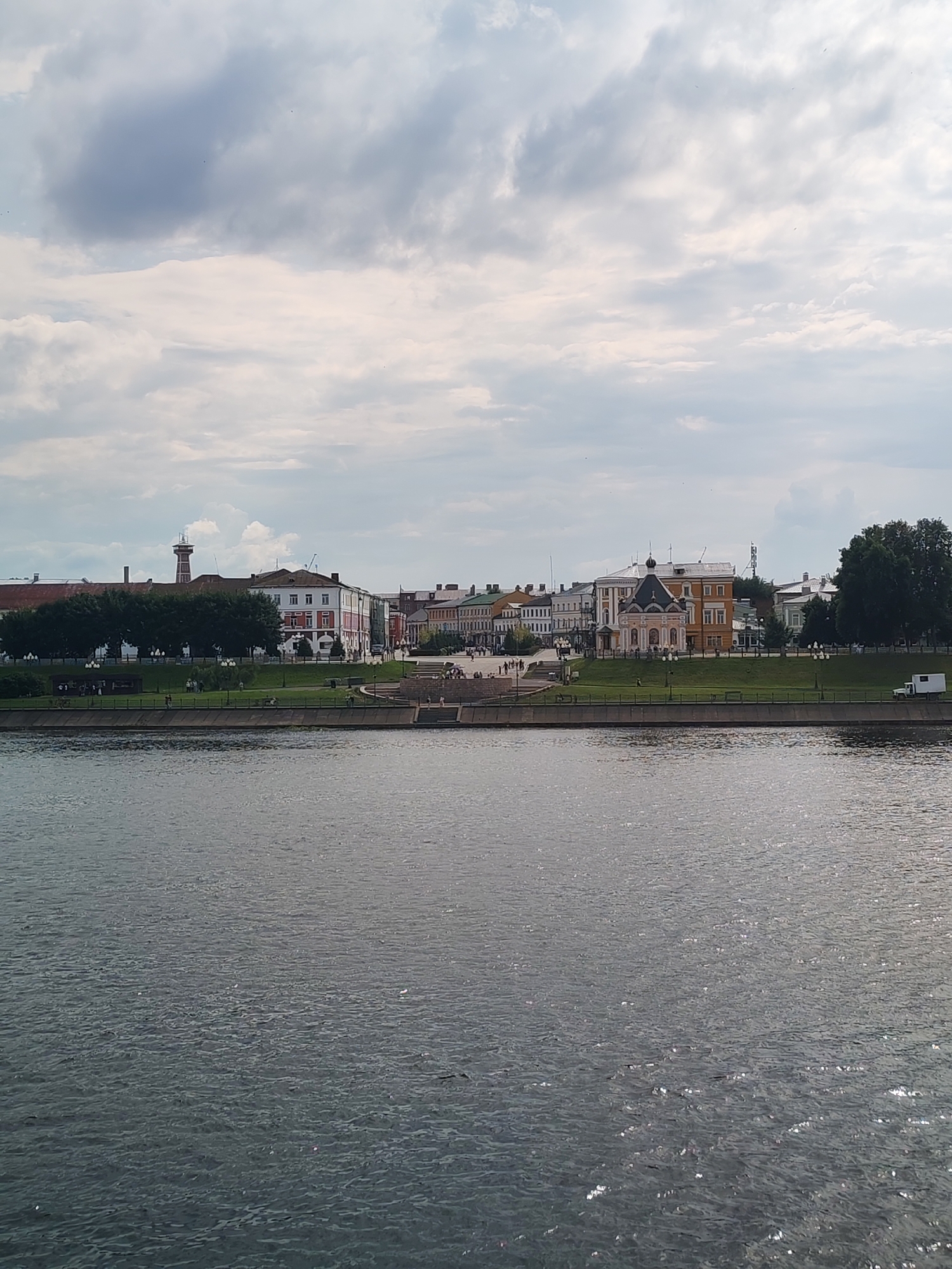 The image depicts a cityscape along a body of water, likely a river, under a cloudy sky. In the background, a line of buildings with various architectural styles, including what appears to be a church with a pink facade and a domed roof, and a tower-like structure resembling a fire watchtower, are visible. Green trees and manicured lawns form a park-like area between the buildings and the waterfront. A concrete embankment with steps leads down to the water, and several people are present on the embankment and in the park area. The water in the foreground is dark and rippled, reflecting the overcast sky. The scene suggests a tranquil urban environment in Bol'shie Mkhi, Russia.