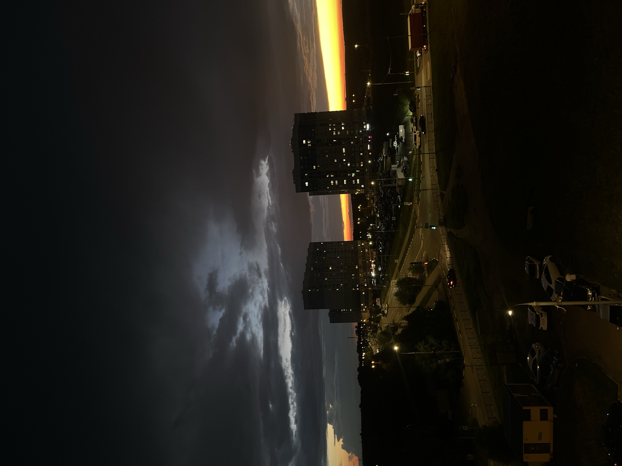 The image depicts a twilight scene in Domodedovo, Russia. The sky is a dramatic blend of dark clouds on the left, gradually transitioning to a vibrant orange and yellow band near the horizon on the right, suggesting sunset or sunrise. Silhouetted against the colorful sky are two multi-story apartment buildings with numerous illuminated windows, indicating that it is evening and people are home.
Below the buildings, a road is visible with streetlights casting a warm glow. Several parked cars are scattered along the roadside and in a parking lot. A large red truck is parked near the top right of the frame. The overall setting appears to be a residential urban or suburban area. The time of day is clearly either dusk or dawn, characterized by the dramatic sky colors and the lit windows in the buildings. The weather appears to be transitioning, with dark, possibly stormy, clouds present alongside the clear, bright horizon. No people are clearly discernible in the image. No visible text is present.