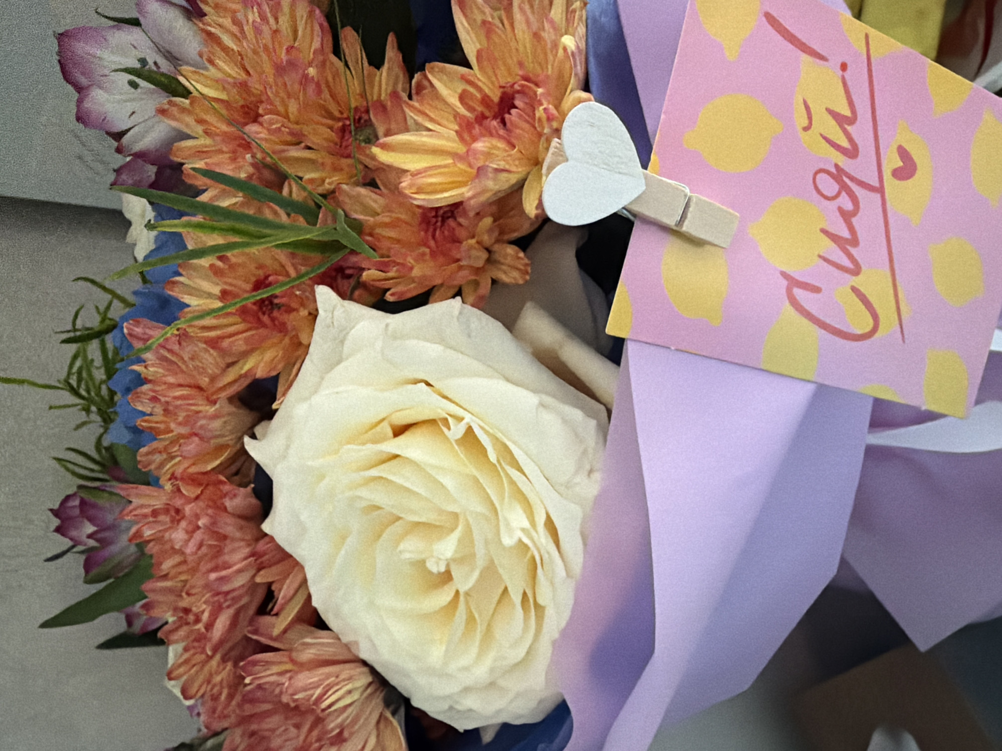 A close-up shot focuses on a bouquet of flowers and a small card attached with a wooden clothespin. The bouquet contains a large, creamy white rose in the center, surrounded by vibrant orange and pink chrysanthemums. Hints of blue and purple flowers are visible in the background, along with green foliage. The flowers are wrapped in light purple paper. A small, pink card with a pattern of yellow lemons is pinned to the wrapping. Handwritten in red script on the card are the Cyrillic letters "Сидя!". A small pink heart is also drawn on the card. The overall scene is well-lit and appears to be indoors. There is no information or visible elements within the image that would suggest a specific location within the city of Petropavlovsk-Kamchatsky, Russia.
