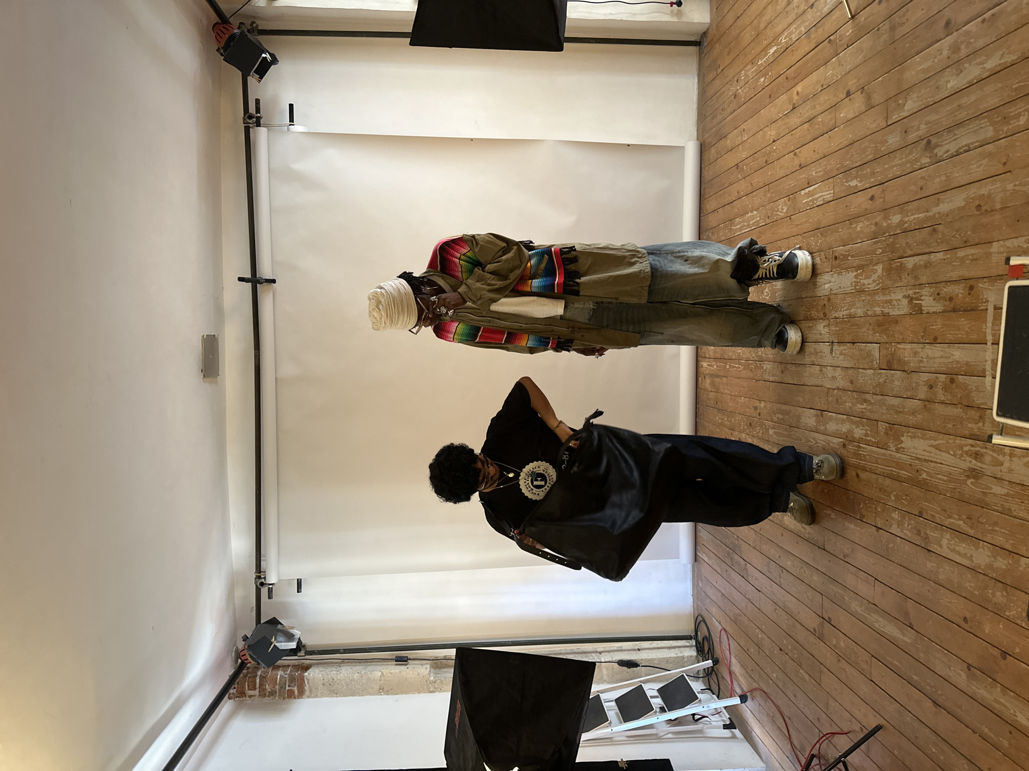 This image depicts an indoor scene with two individuals standing in what appears to be a photography studio in Paris, France, suggested by the style of the exposed brick on the lower left side of the photo. The studio features a white backdrop with a wooden floor.
One individual is wearing a turban, glasses, a multi-colored jacket, and jeans. They are standing upright.
The other individual is wearing a black shirt and black pants, and is holding a large black bag. This person is looking down and appears to be in motion.
Lighting equipment is visible, including softboxes and lighting stands, which confirms the location as a photography studio. A small stepladder is also present in the foreground. The scene appears to be during a photo shoot or setup.