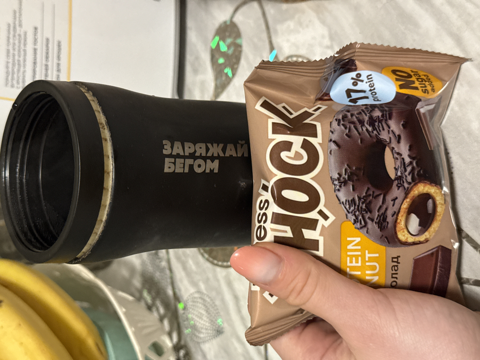 A hand holds a package of "ess HOCK" protein donut in front of a black thermos bottle with the words "ЗАPЯЖАЙ БЕГОМ" (CHARGE RUN) printed on it. The package advertises "17% protein" and "NO sugar added." The background includes a blurred surface with a leaf-like pattern, and a partially visible bowl of bananas in the lower left. The scene is indoors. The precise location in Chaikovsky, Russia cannot be determined from the image.