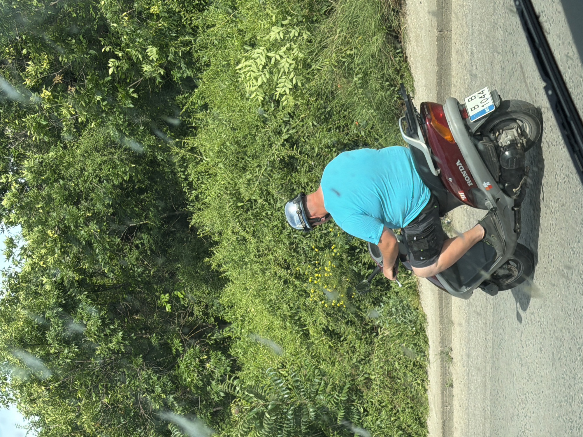 A person wearing a light blue t-shirt and black shorts rides a red and grey Honda scooter on a paved road. The person is wearing a helmet. The scooter has a license plate that reads "BG 8 1044". The scene is outdoors with abundant green foliage on the left side of the road, suggesting a rural or suburban setting. The road appears to be a two-lane road with a concrete curb on the left. The weather appears to be sunny, as indicated by the bright lighting. The image is taken from a slightly elevated angle, possibly from another vehicle. There are some light streaks or smudges on the lens or window from which the photo is taken.