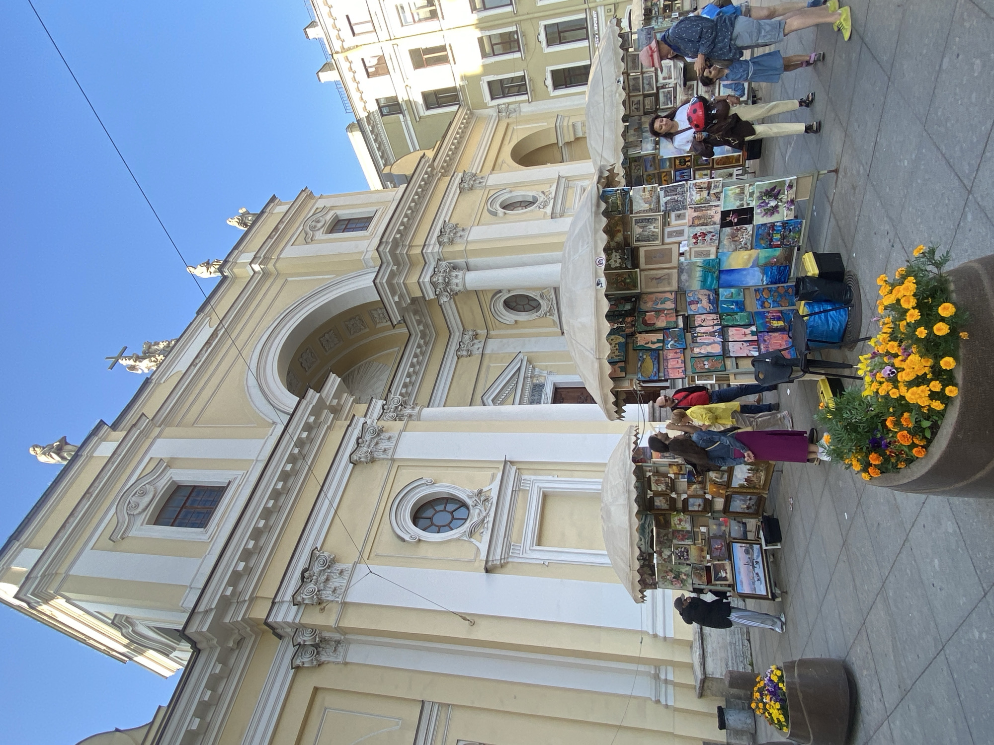 This is an outdoor scene in Saint Petersburg, Russia, likely on a sunny day, showing a street art market in front of a light yellow building with classical architectural elements, possibly a church. Several people are browsing or standing near stalls displaying numerous paintings and artworks. One stall has a white umbrella canopy, and the paintings are arranged on stands, showcasing a variety of styles and subjects, including portraits, landscapes, and still lifes. In the foreground, there are two large planters filled with vibrant orange and yellow flowers. A few individuals are visible examining the art, with some in conversation. The building in the background features arched entrances, columns, ornate window surrounds, and statues on its roofline. The ground is paved with grey tiles.