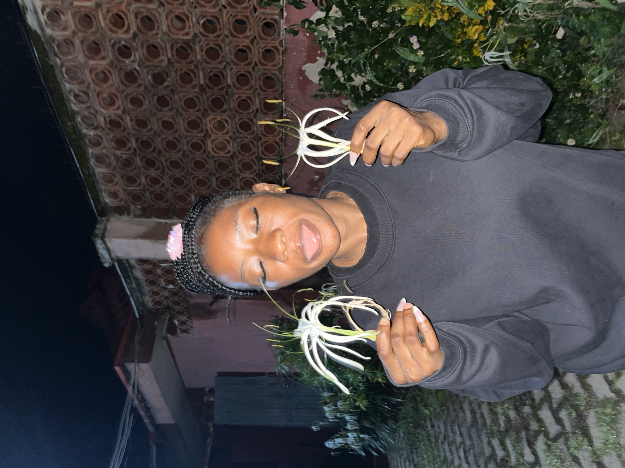 A young Black woman with dark braided hair, a pink hair accessory, and closed eyes is shown from the chest up, sticking her tongue out. She is wearing a black crewneck sweatshirt and gold earrings. She is holding a white, spidery flower in each hand. The background shows a textured, reddish-brown wall, a pink building, and lush green foliage. The ground is paved with interlocking gray stones. The scene appears to be outdoors at night, possibly in Jos, Nigeria, given the architectural style of the wall and building.