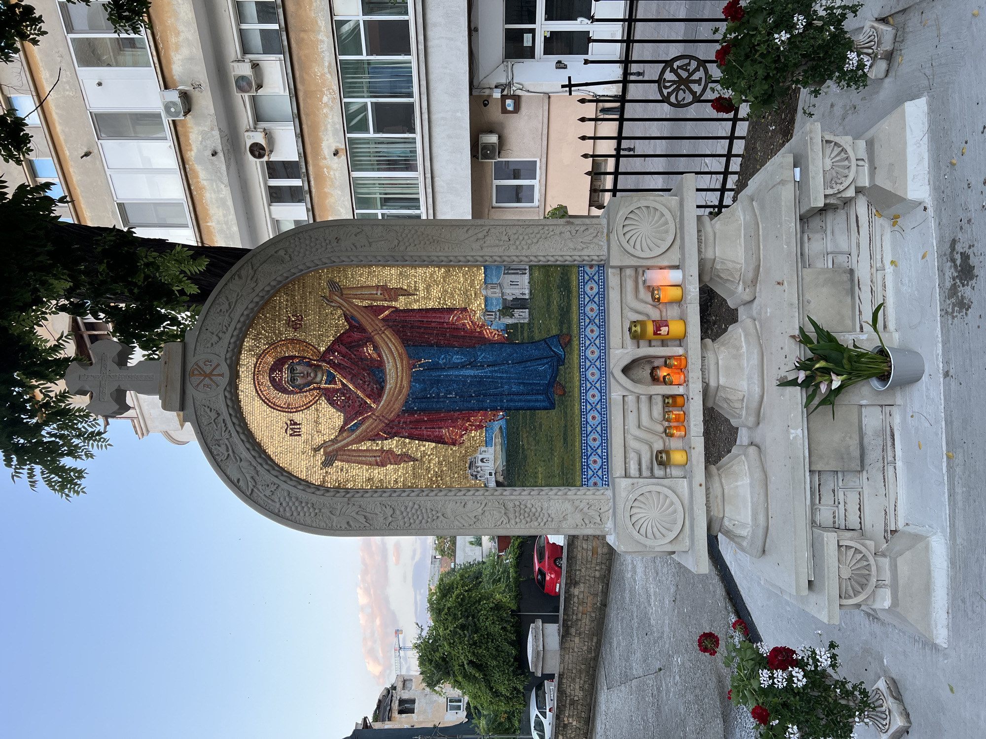 A religious mosaic icon of the Virgin Mary, rendered in gold and vibrant colors, is displayed outdoors on a stone monument in Constanța, Romania. The icon is framed by an arched structure adorned with decorative carvings and a cross at its apex. Below the mosaic, several votive candles are lit, casting a warm glow. The monument is situated on a paved area, with a metal fence behind it and a building in the background. Red and white flowers in planters flank the monument. The sky is a soft blue with hints of pink clouds, suggesting it is either early morning or late afternoon.