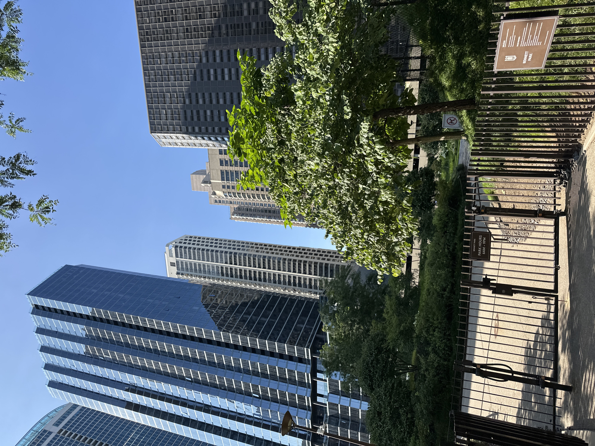 The image depicts a view of Bennett Park in Chicago, Illinois, USA. In the background, several tall modern skyscrapers dominate the skyline, with one prominent building featuring a dark blue glass facade. In the foreground, a black metal fence encloses the park, with a gate in the center. A sign attached to the fence indicates "BENNETT PARK PARK HOURS 6AM - 9PM". Lush green trees and landscaping are visible within and around the park, contrasting with the urban architecture. The sky is clear and blue, suggesting a sunny day. There are no people visible in the image.