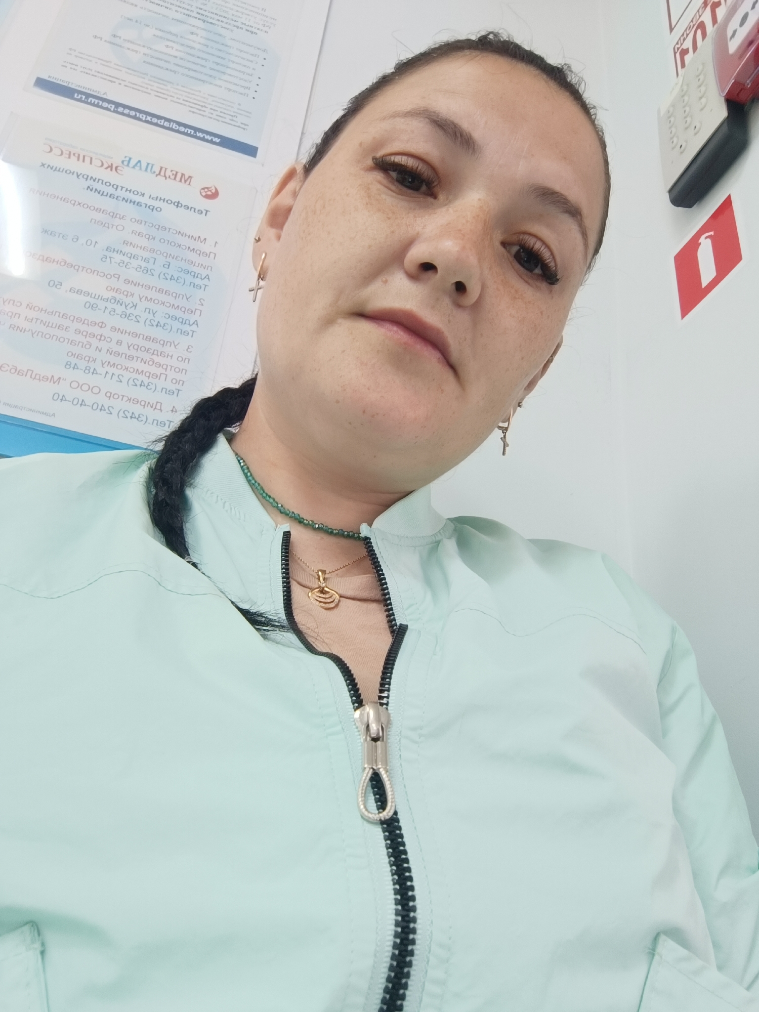A medium shot of a woman with dark hair and freckles on her face, wearing a light mint green zippered jacket over a tan shirt, is captured from a slightly high angle. She has on a gold pendant necklace and a beaded green necklace. A cross-shaped earring is visible on her left ear. The background shows a white wall with some signs and a fire alarm system mounted on it. The overall lighting is bright, suggesting an indoor setting. Based on the visible text in Russian on documents in the background, specifically mentioning "Чайковский" (Chaikovsky), the location is likely within the city of Chaikovsky, Russia.