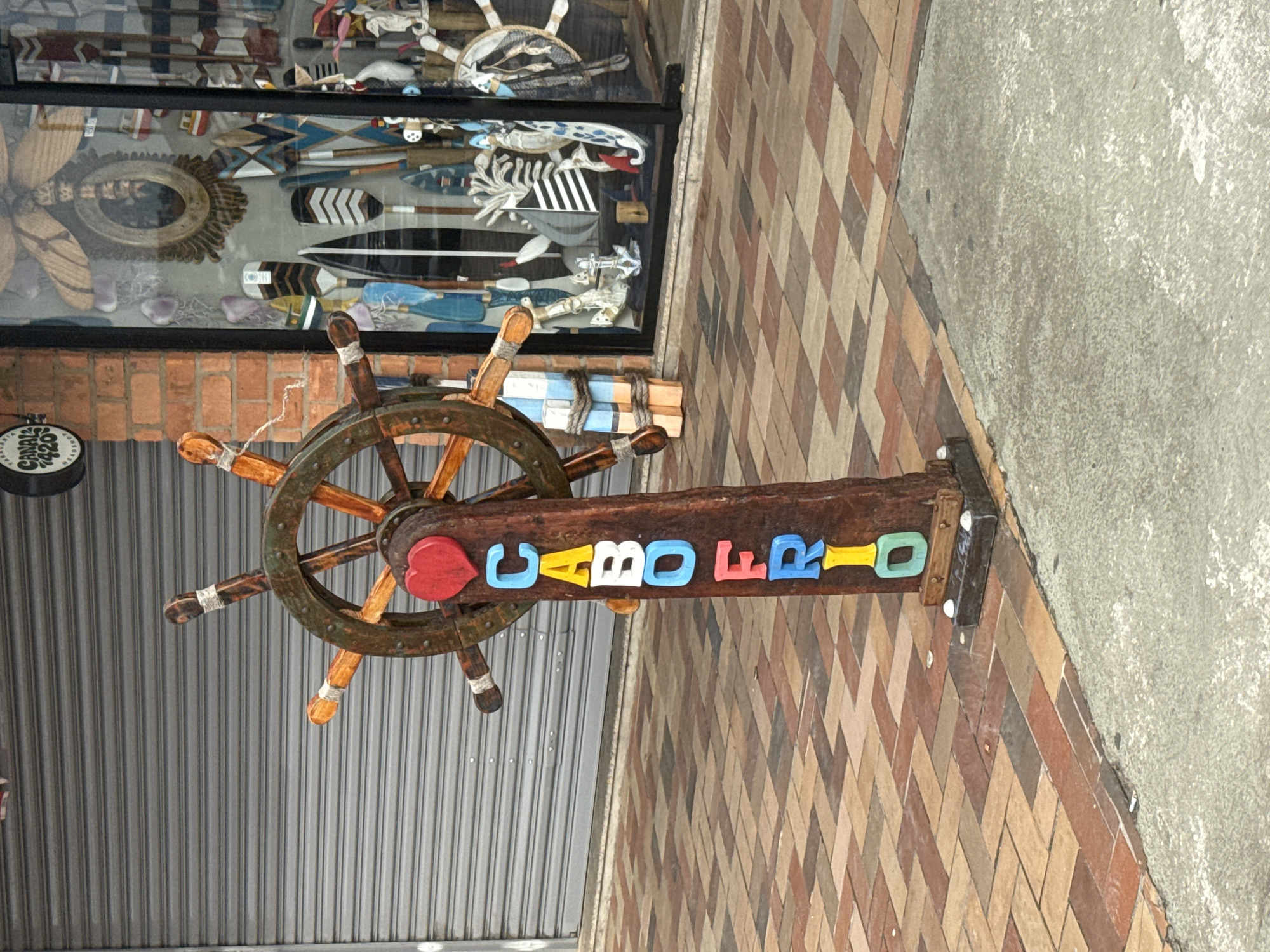 This is a medium shot, eye-level, outdoor photograph taken in Cabo Frio, Brazil. The image features a wooden post adorned with colorful, three-dimensional letters spelling out "CABO FRIO." A red heart sits at the top of the post, directly above the letters. The post is a part of a nautical-themed decoration, as it is topped with a ship's steering wheel made of weathered wood. The steering wheel has eight spokes and is partially visible in the upper portion of the frame.
The background includes a gray, corrugated metal roll-up door on the left, and a shop window on the right. The shop window displays a variety of nautical-themed items, including oars, decorative paddles, anchors, and a miniature sailboat. The foreground shows a sidewalk paved with reddish-brown and tan square tiles, leading to a gray concrete area. The overall scene suggests a commercial area, likely a street in the city of Cabo Frio, perhaps near a souvenir shop or a business with a maritime theme. There are no people visible in the image.