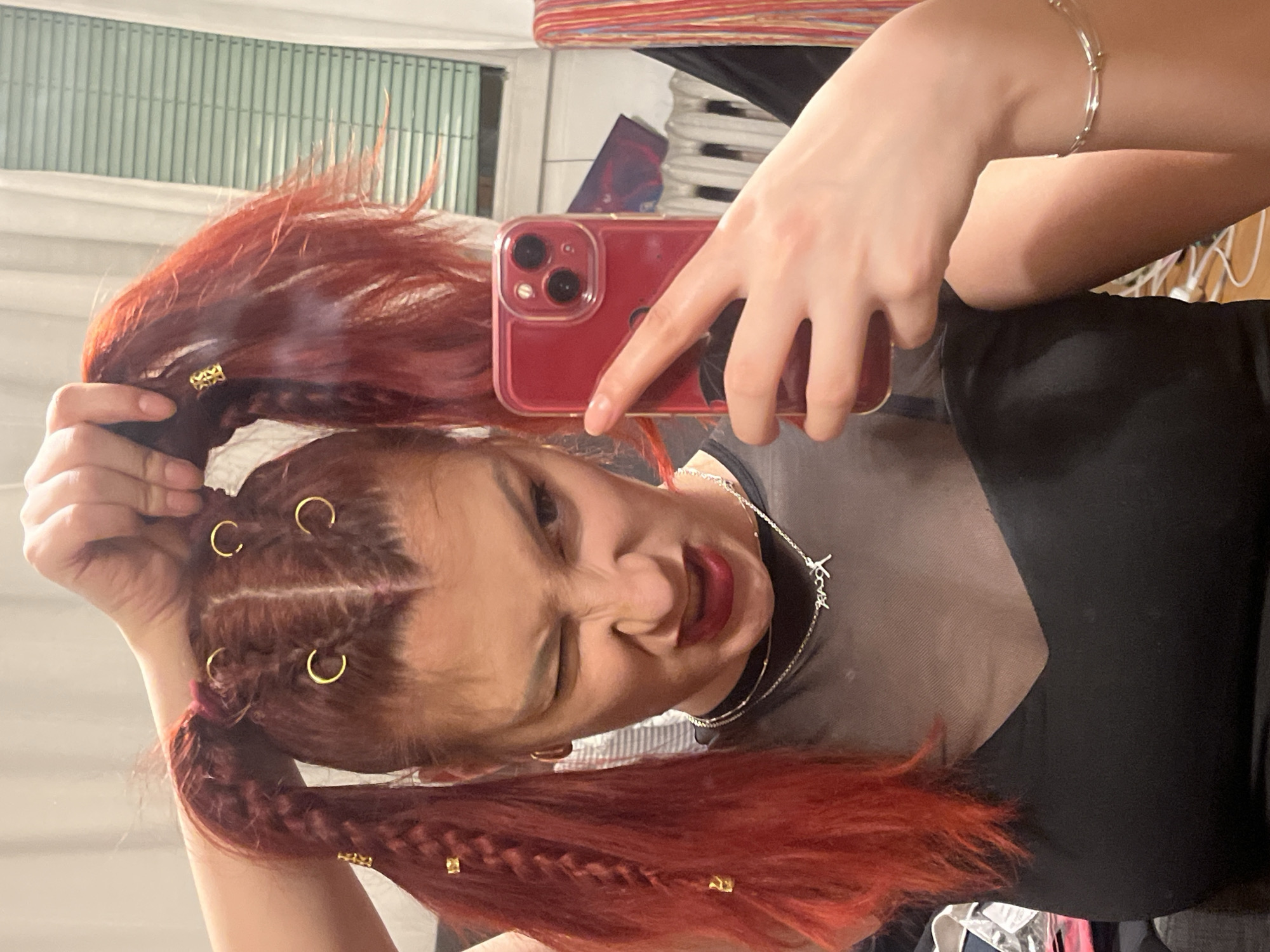 A person with red braided hair is taking a selfie. The person is holding a red phone and wearing a black top and silver jewelry. The location is not discernible.