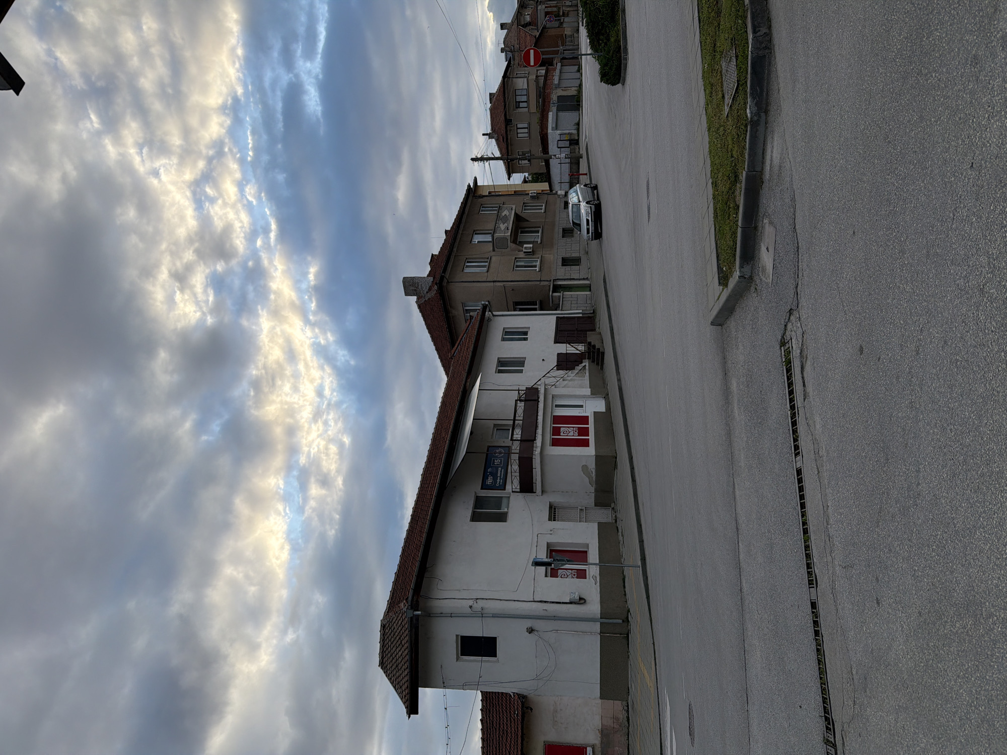 The image depicts a street scene in Panagyurishte, Bulgaria. The foreground features a gray asphalt road with a drainage grate running along its side. Adjacent to the road is a sidewalk with a patch of greenery.
On the left, a multi-story building with a white facade and a red-tiled roof stands prominently. The building has a series of windows, some of which are visible, and at least two balconies with red accents. Signage in a local language is visible on the building's facade.
Further down the street, another multi-story building is visible, its facade a darker gray color. A parked silver car is positioned near the base of this building.
In the background, additional buildings with red-tiled roofs and varying architectural styles can be seen. A "do not enter" road sign is also present.
The sky overhead is filled with dramatic clouds, with some areas reflecting sunlight.