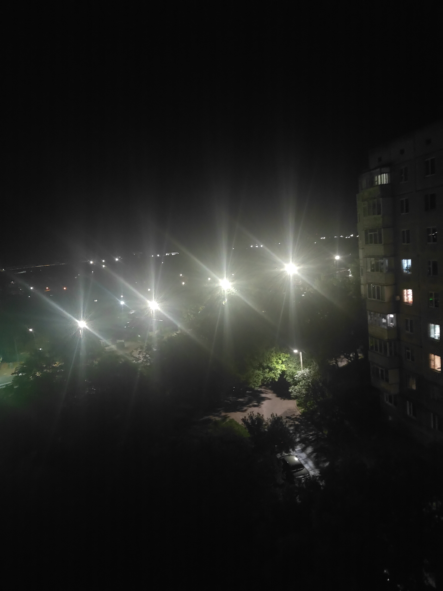 The image depicts a night view of an urban residential area in Bila Tserkva, Ukraine. The foreground is dominated by the side of a multi-story apartment building. The building appears to be constructed of concrete panels, with numerous windows, some of which are illuminated. Below and to the left of the building, there is a dark area with trees and a road. Further into the scene, multiple streetlights with distinct lens flares illuminate a street and surrounding park-like areas with trees. In the background, distant lights suggest a more densely populated part of the city or a highway. The overall impression is one of a quiet night in a typical Eastern European residential neighborhood.