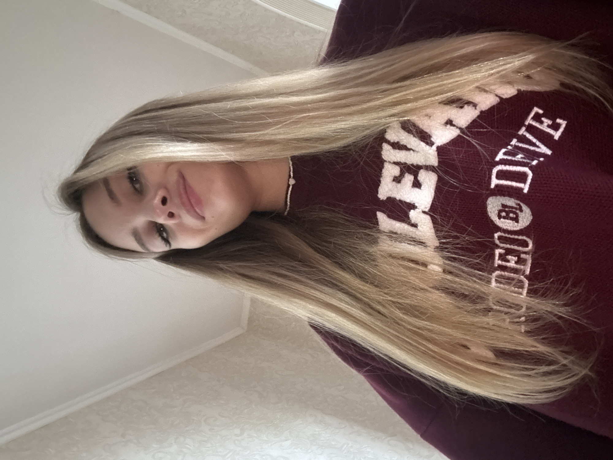 This is a close-up, low-angle selfie of a young woman with long, blonde hair. She is wearing a dark red sweater with white lettering. The visible text on the sweater reads "LEVE BJODE." She has light skin, dark eyeliner, and a slight smile. A delicate beaded necklace with a small heart pendant is visible around her neck. The background appears to be an indoor setting with a white ceiling and patterned wallpaper. The lighting suggests it is daytime. The overall impression is casual and personal.