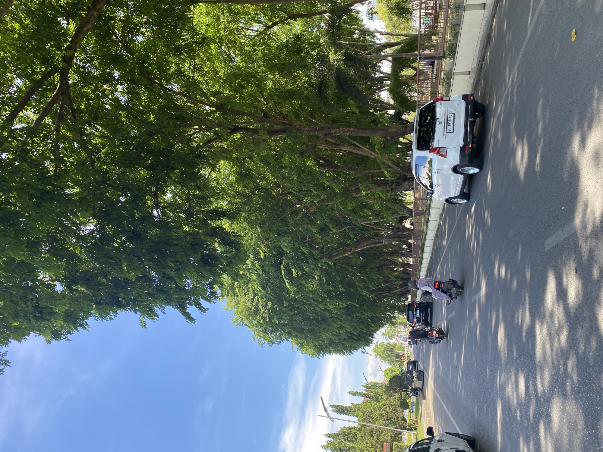The image depicts a street scene in the city of Lhokseumawe, Indonesia, on a clear, sunny day. A paved road runs through the center of the frame, lined on the right by a dense row of mature trees with lush green foliage, casting dappled shadows onto the asphalt. Beyond the trees, a brown metal fence separates the road from a walkway or park area.
Several vehicles are present on the road. A white SUV, possibly a Toyota Avanza based on its design and the license plate "BL 1518 AJ," is parked on the right side of the road. In front of it, two motorcycles are traveling away from the camera. The rider of the motorcycle closest to the SUV is wearing a white headscarf and a light purple outfit. The rider of the second motorcycle, further ahead and to the left, is wearing a dark outfit and a helmet. A black SUV is also visible further up the road, behind the motorcycles. On the far left, a portion of a white car is visible, facing away from the camera.
The left side of the road features a sidewalk, a few smaller trees, and some buildings in the distance. Tall, slender lamp posts are also visible. The sky is a bright blue with scattered white clouds. The overall impression is of a well-maintained urban street with significant tree cover, indicative of a tropical climate.