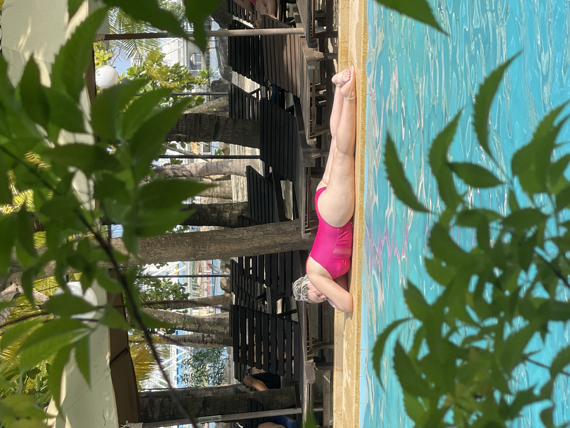 A woman in a pink swimsuit is reclining poolside in what appears to be the Tay Natural Heritage Area Trang, Vietnam. She is positioned next to a turquoise swimming pool, with lounge chairs arranged nearby. Palm trees are visible in the background and foreground, adding to the tropical environment. The scene is shot through foliage, partially obscuring the view. Other people are partially visible sitting on the chairs.