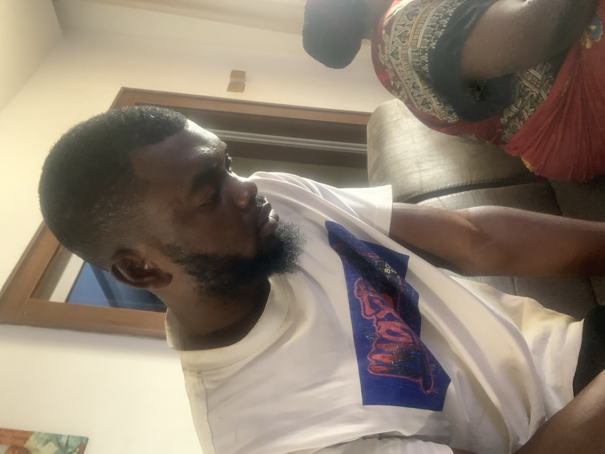 An indoor, horizontally oriented shot captures two individuals in a relaxed setting, likely a living room in Jos, Nigeria. The main subject, a Black man, is featured prominently in the foreground, seen in profile looking towards the viewer's left. He has a close-cropped haircut and a dark, full beard. He is wearing a white crew-neck t-shirt adorned with a large blue graphic design on his chest, which includes stylized, unreadable text in red and white.
Behind him and to his right, a second person, also appearing to be Black, is partially visible from the upper back and shoulder. This individual is wearing a distinctive garment with a patterned fabric in shades of red, black, and cream or gold, possibly a traditional attire. Both individuals appear to be seated on a light-colored, upholstered sofa, part of which is visible in the background.
The room features light-colored walls and a ceiling. A wooden door or window frame is visible on the left side of the image, reflecting some light. A small, framed piece of artwork is partially visible in the bottom-left corner. The lighting is soft, suggesting an indoor setting without direct sunlight.