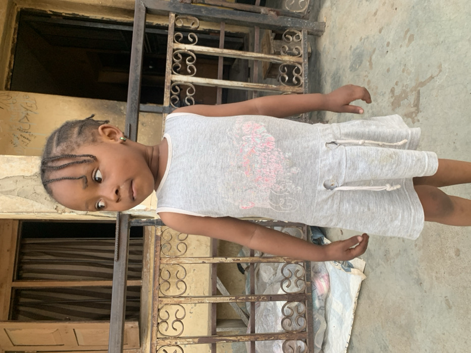 A young, dark-skinned girl stands in front of an old, weathered building with decorative wrought iron railings. She wears a light grey dress with a faded design and has braided hair. Her expression is curious, as she looks off to the side. The background features a combination of textured walls, dark doorways, and a glimpse of a white material on the ground. The image appears to be taken outdoors in daylight, with a slight tilt.