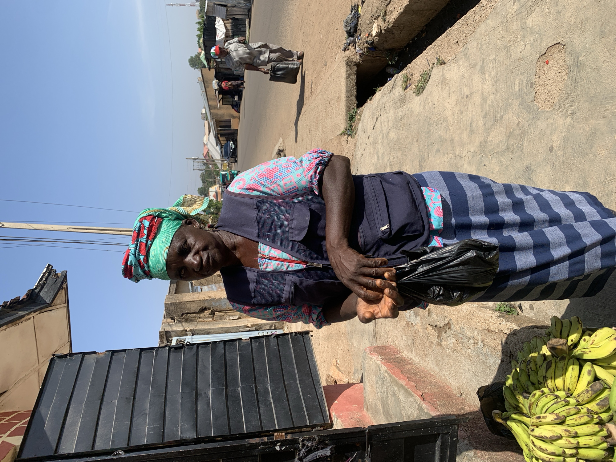 An elderly African woman stands on a concrete sidewalk in Jos, Nigeria. She wears a blue and pink patterned top under a dark blue vest, a blue and gray striped skirt, and a colorful headscarf. She holds a black plastic bag. To her right, a pile of green and yellow bananas rests on the ground. To her left is a black metal gate. In the background, another person walks down the street. The scene appears to be an urban environment with buildings and infrastructure visible.