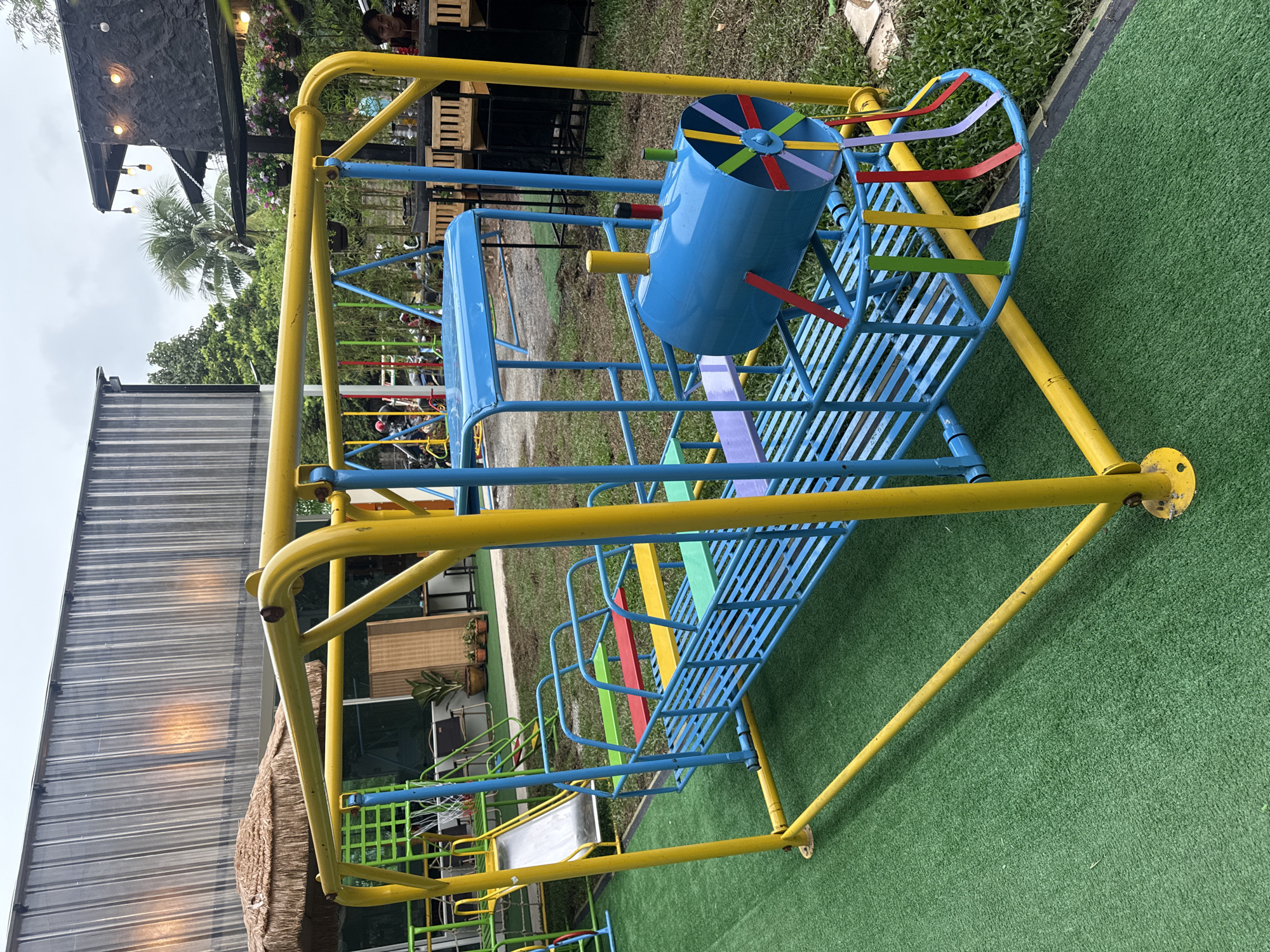 The image shows a blue and yellow playground structure resembling a train, located outdoors on a green artificial turf surface. The structure has multiple seats colored red, yellow, green, and purple. A blue cylindrical object with colorful ribbons emanating from it is attached to the front of the train, likely representing a smokestack or engine part. In the background, there is a building with a corrugated metal roof, a thatched umbrella, trees, and some outdoor seating with patrons. The location is identified as a playground in Wichit, Thailand.