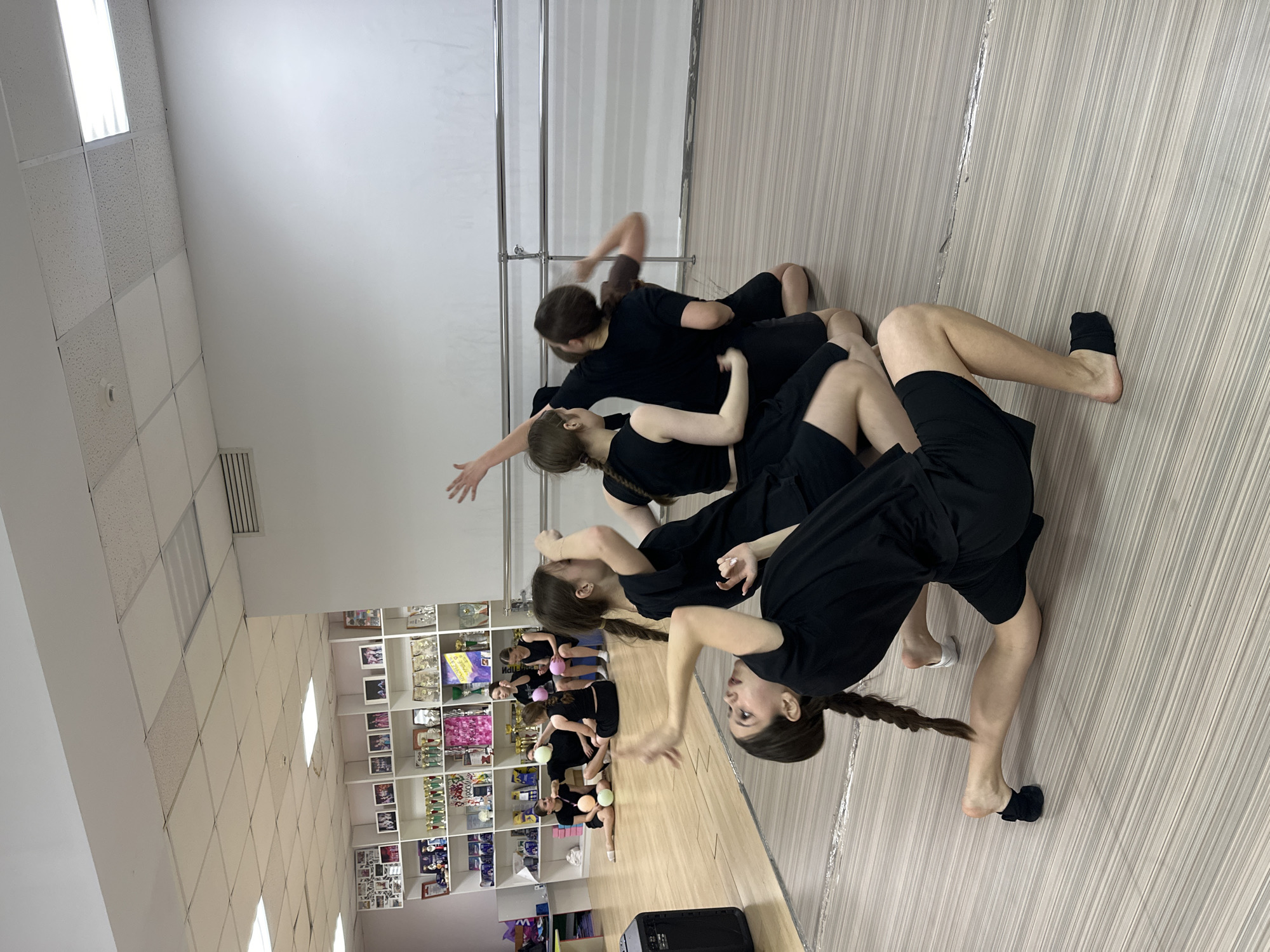 The image captures a group of approximately 7-8 young individuals, predominantly female, engaged in a dance practice within a studio. They are dressed in matching black athletic wear, consisting of t-shirts and shorts, with some wearing black or white dance socks, while others are barefoot. The subjects are positioned in various synchronized and artistic poses, some appearing to be stretching, balancing, or performing contemporary dance movements. Their expressions suggest focus and concentration.
The setting is an indoor dance studio featuring a light-colored wooden floor, white walls, and a suspended ceiling with square acoustic tiles and integrated fluorescent lighting. A large mirror along one wall reflects parts of the room and additional dancers, some of whom are seen seated on the floor with small, colorful exercise balls (pink, green, yellow), suggesting warm-up or conditioning exercises. A silver dance barre is partially visible near the mirror.
On the left side of the room, a white shelving unit is filled with numerous trophies, awards, framed pictures, and other memorabilia, indicating a history of achievements and a dedicated dance facility. A black portable speaker rests on the floor in the foreground. The lighting is bright, suggesting daytime. Minimal visible text includes a faint Cyrillic word, possibly "МОЛОДЕЦ" (Molodets, meaning "Well done"), on one of the awards. The scene reflects a diligent training session in a dance school in Votkinsk, Russia.