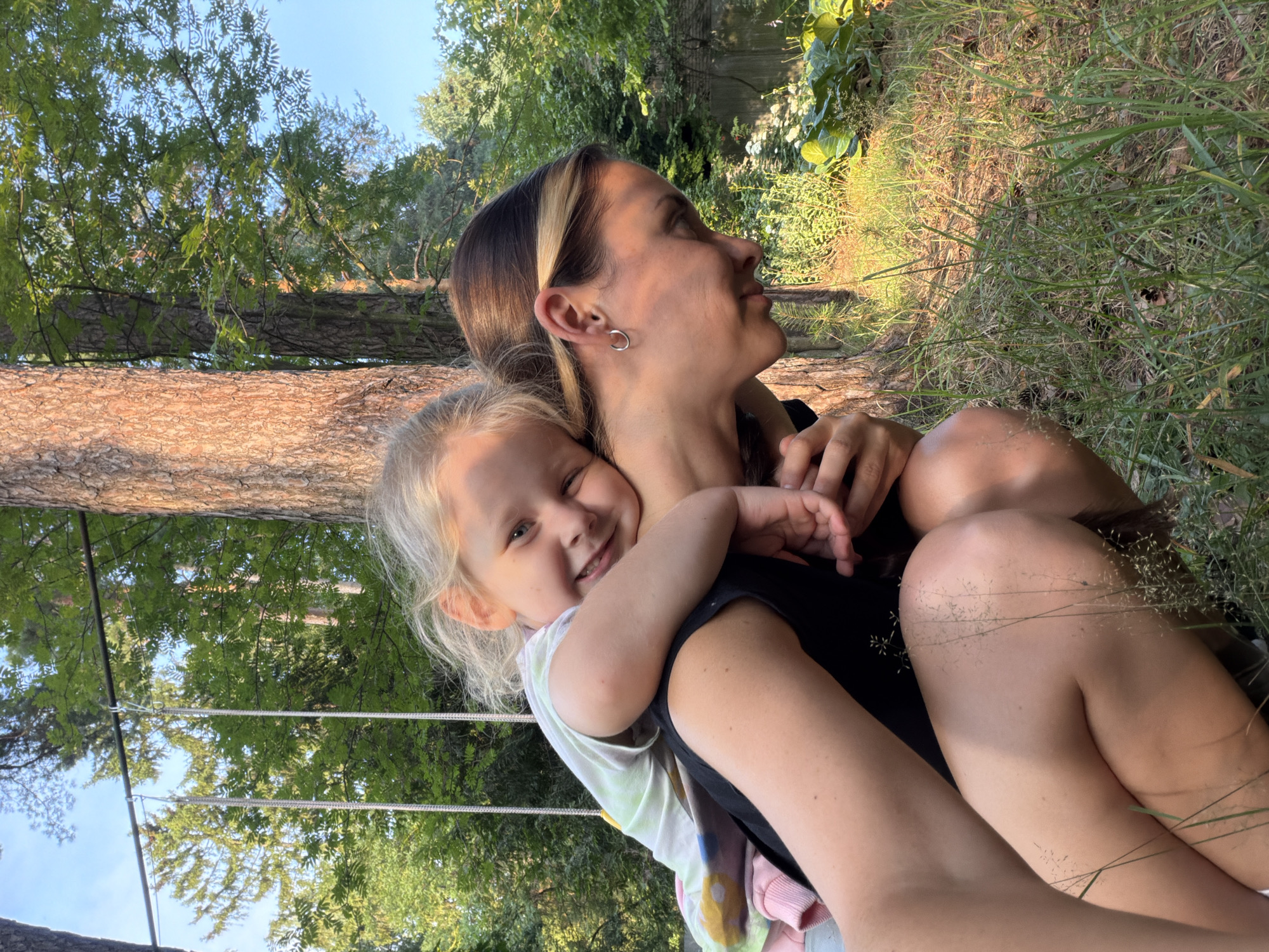In this candid outdoor photograph, a woman and a young child are embracing, seated amidst a natural setting. The woman, with blonde-streaked brown hair and a hoop earring, looks upwards and to the right with a gentle expression. The child, with blonde hair, is nestled in the woman's arms, smiling broadly at the camera and hugging the woman's neck. Both are dressed casually; the child wears a tie-dye shirt, and the woman a black top.
The background suggests a park or wooded area in Udelnaya, Russia. A large tree trunk, with rough bark, is prominent to the left, and lush green foliage and trees form the backdrop. The scene appears to be lit by natural sunlight, indicating it's likely daytime, possibly late afternoon given the warm, golden quality of the light filtering through the trees and illuminating the grass. The ground is covered with dry grass and some scattered dry seed heads. In the upper left quadrant of the image, a portion of what appears to be a hammock or swing set with ropes and metal supports is visible against a clear blue sky. The overall mood is one of warmth, affection, and peaceful enjoyment of nature. There is no visible text in the image.