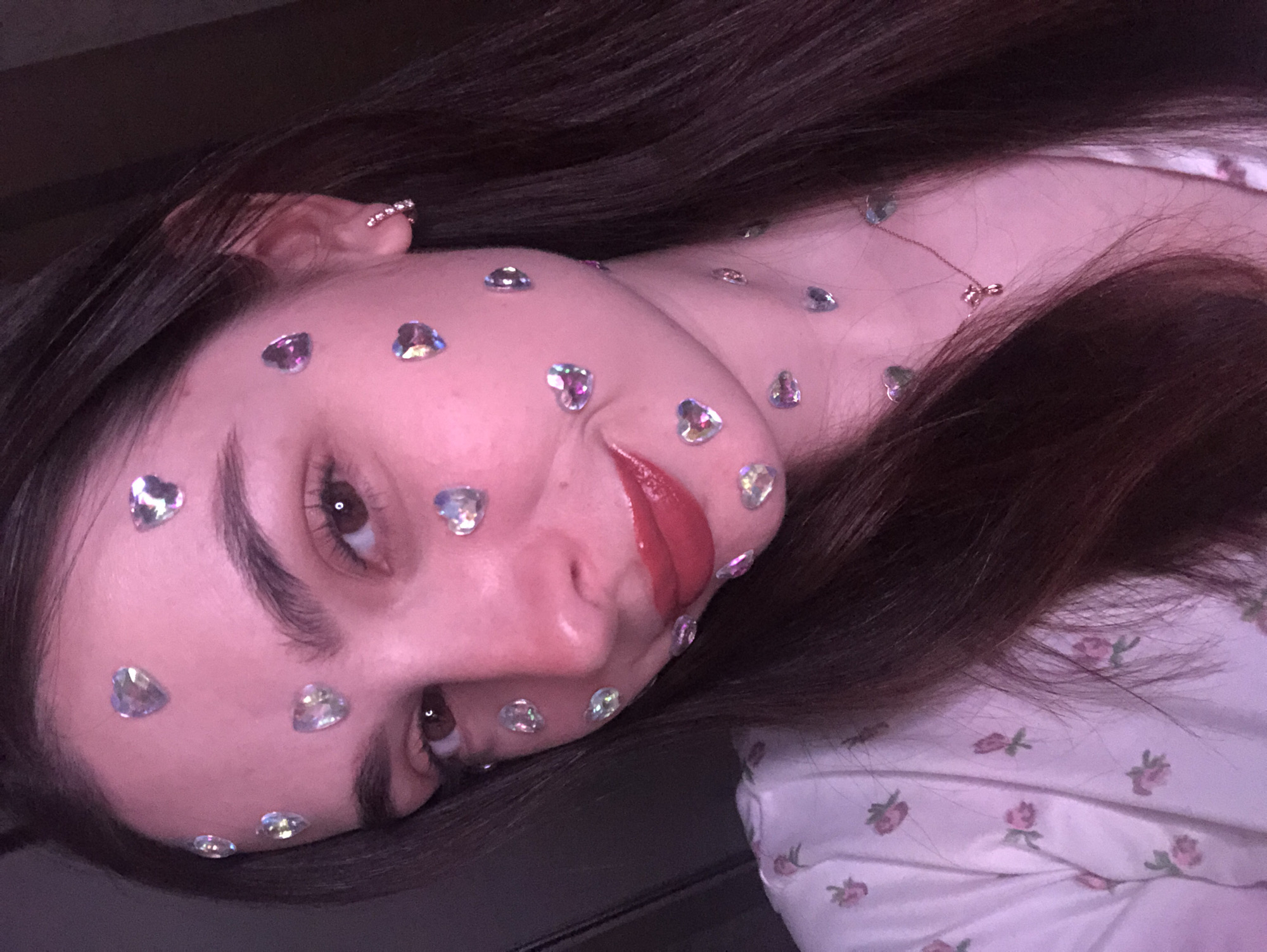 A young woman with long, dark brown hair is lying on a dark surface, possibly a bed or sofa. She is wearing a white top with a pink floral pattern. Her face and neck are decorated with small, iridescent heart-shaped stickers. She has brown eyes and appears to be wearing lip gloss. A small earring or ear cuff is visible on her left ear. A delicate necklace with a bow-shaped pendant is visible on her neck. The lighting is soft and appears to be coming from above. The background is dark and indistinct. There is no indication of the location being Noviy, Russia.