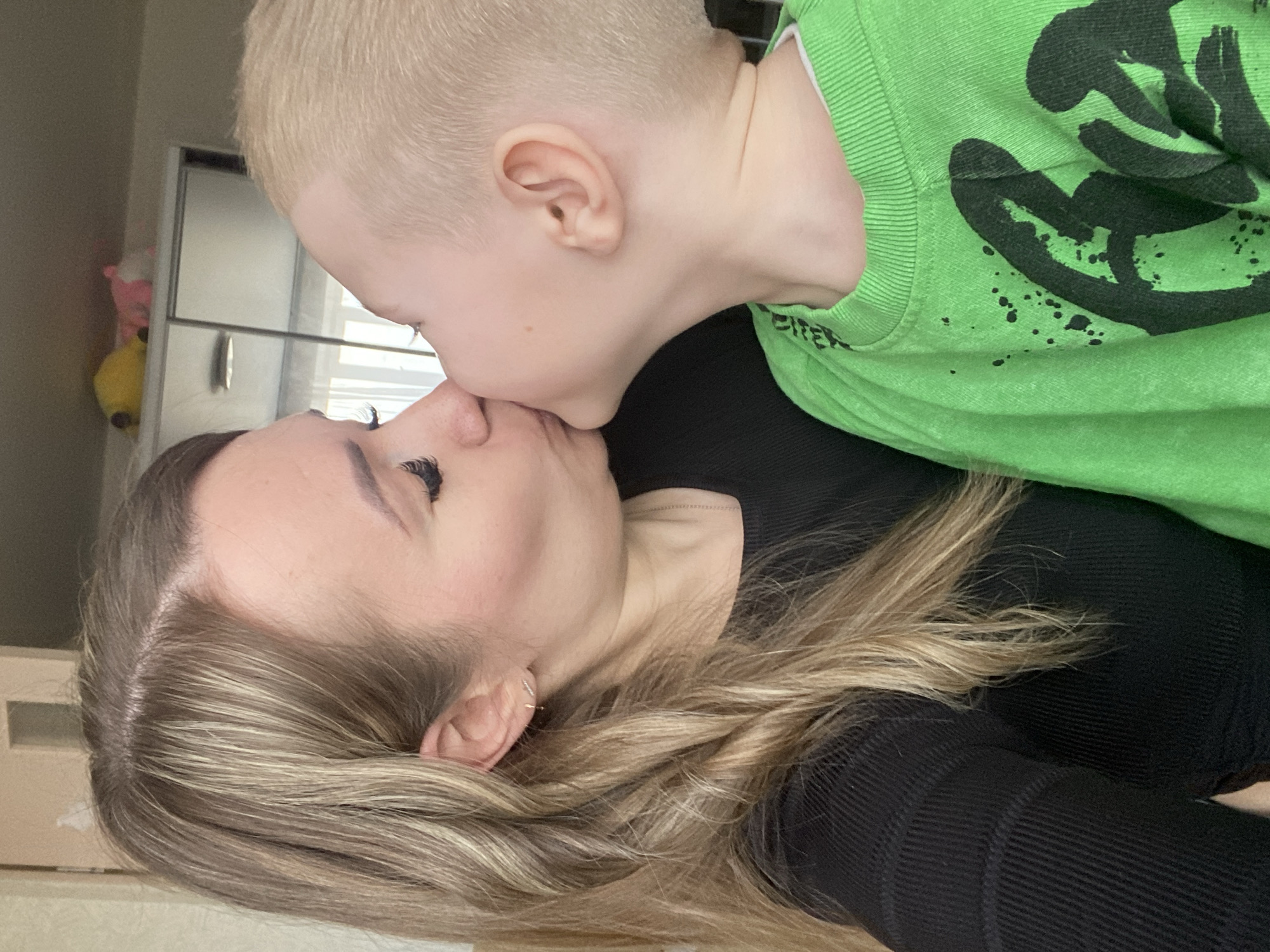 A fair-skinned woman and a fair-skinned child are closely embracing. The woman lies on her back, wearing a black long-sleeved shirt, with blonde highlighted hair framing her face. She has an earring visible in her left ear. The child, wearing a bright green sweatshirt with a black design, is positioned above her and giving her a kiss on the nose. The background reveals a room interior, possibly a home, with a blurred view of a white cabinet or refrigerator, as well as blinds visible behind the cabinet. There are a few stuffed animals visible out of focus on top of the cabinet. The location is not possible to determine based solely on the visual elements in the image and is not in Izhevsk, Russia.