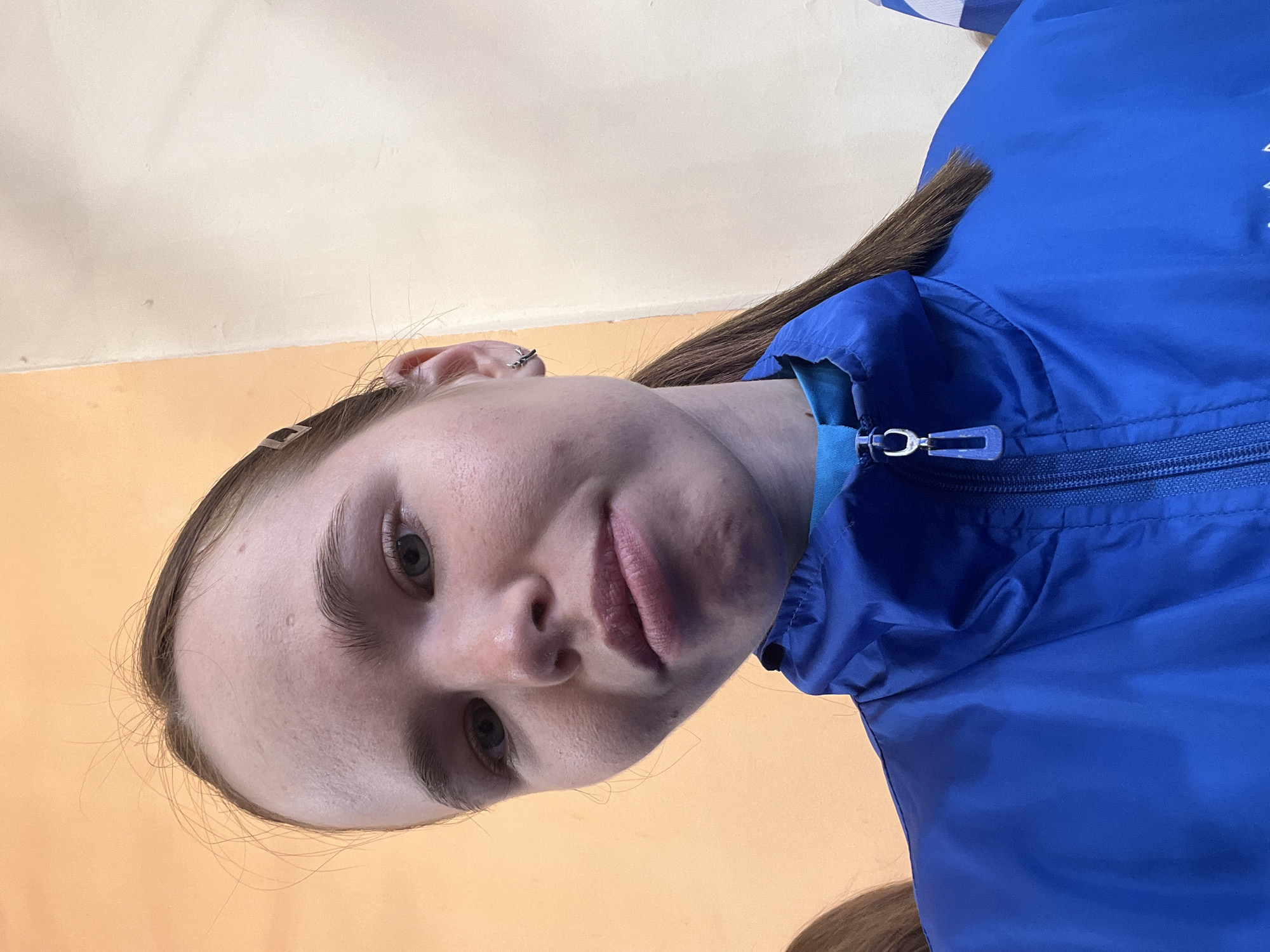 A close-up, angled shot depicts a young woman with fair skin, brown hair, and light-colored eyes. She is wearing a blue jacket with a visible zipper. The background consists of a wall with two color tones, a light yellow/cream color towards the top and a more orange color below. The photograph might have been taken indoors. There is no verifiable evidence within the image to definitively place the location in the city of Chaikovsky, Russia.