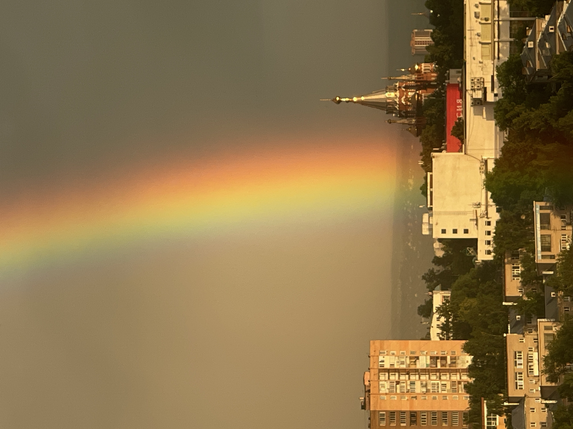 The image shows a city skyline at sunset, dominated by a vibrant rainbow arching across the sky. In the foreground, several multi-story buildings are visible, some with apartment-style balconies. A distinctive red-brick building with multiple onion domes and golden spires, characteristic of Russian Orthodox churches, stands out amongst the cityscape, partially obscured by trees. A large white building with a red sign displaying Cyrillic text is situated in front of the church. The sky is a gradient of warm yellows and muted grays, suggesting lingering daylight and the possibility of recent rain. The overall scene captures a moment of natural beauty within an urban environment.
