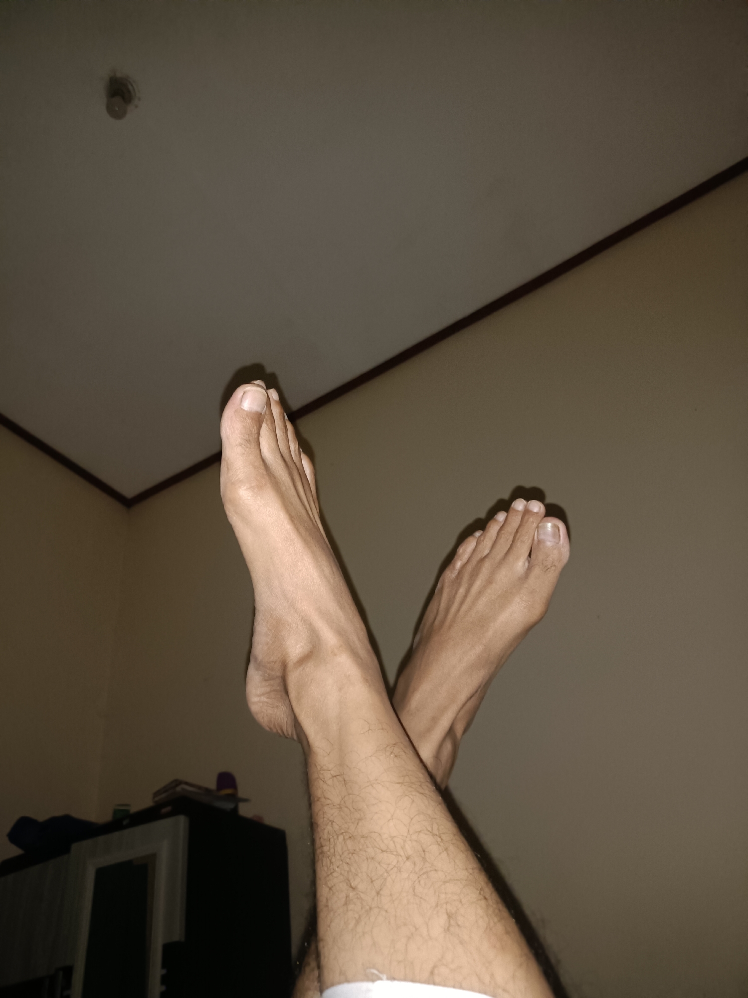 The image shows a low-angle shot of a person's bare feet crossed in front of a wall and ceiling. The feet are facing upwards and outwards, with visible toenails and the contours of the bones and tendons of the feet. The skin appears to be light-skinned, and there is visible hair on the lower legs. The background consists of a plain, light-colored wall and a white ceiling with a thin dark line separating them. A smoke detector is visible on the ceiling. In the lower left of the frame, the top of a dark piece of furniture, possibly a cabinet or dresser, is visible. The lighting is dim, casting shadows on the wall and ceiling. The scene appears to be indoors. There is no indication that this image is located in Kediri City, Indonesia.