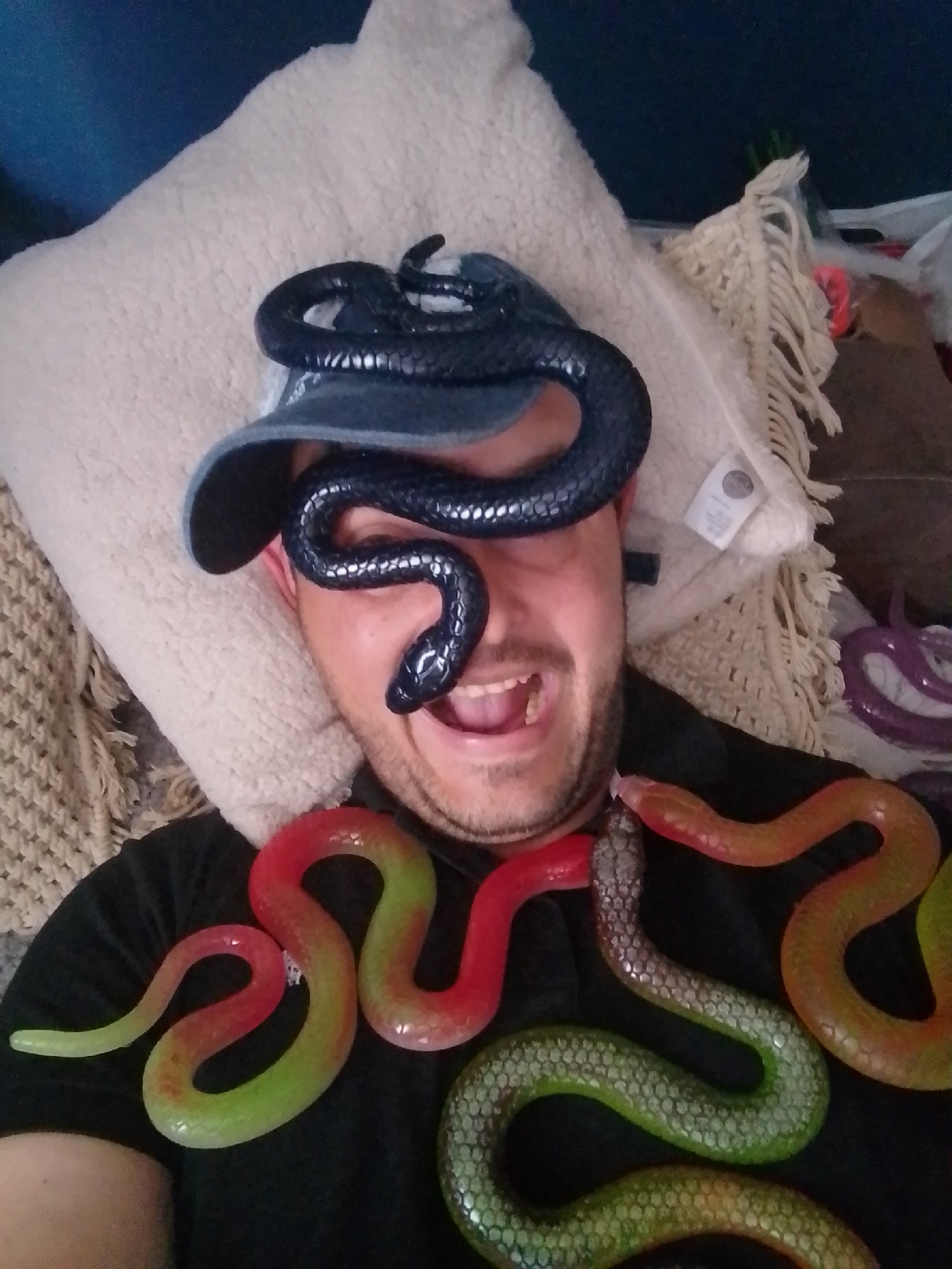 The image shows a man wearing a dark t-shirt and a baseball cap. A black snake toy is draped over his head, with its body covering his eyes and nose. Several other colorful snake toys, red, green, and orange, are placed around him, some on his t-shirt and others in the background. He is smiling and his mouth is open. The man is lying down on a beige textured cushion. The background is dark and indistinct, with some fringed fabric visible to the right. There is no information in the image that allows for the determination of the location in the city of Wedel, Germany.