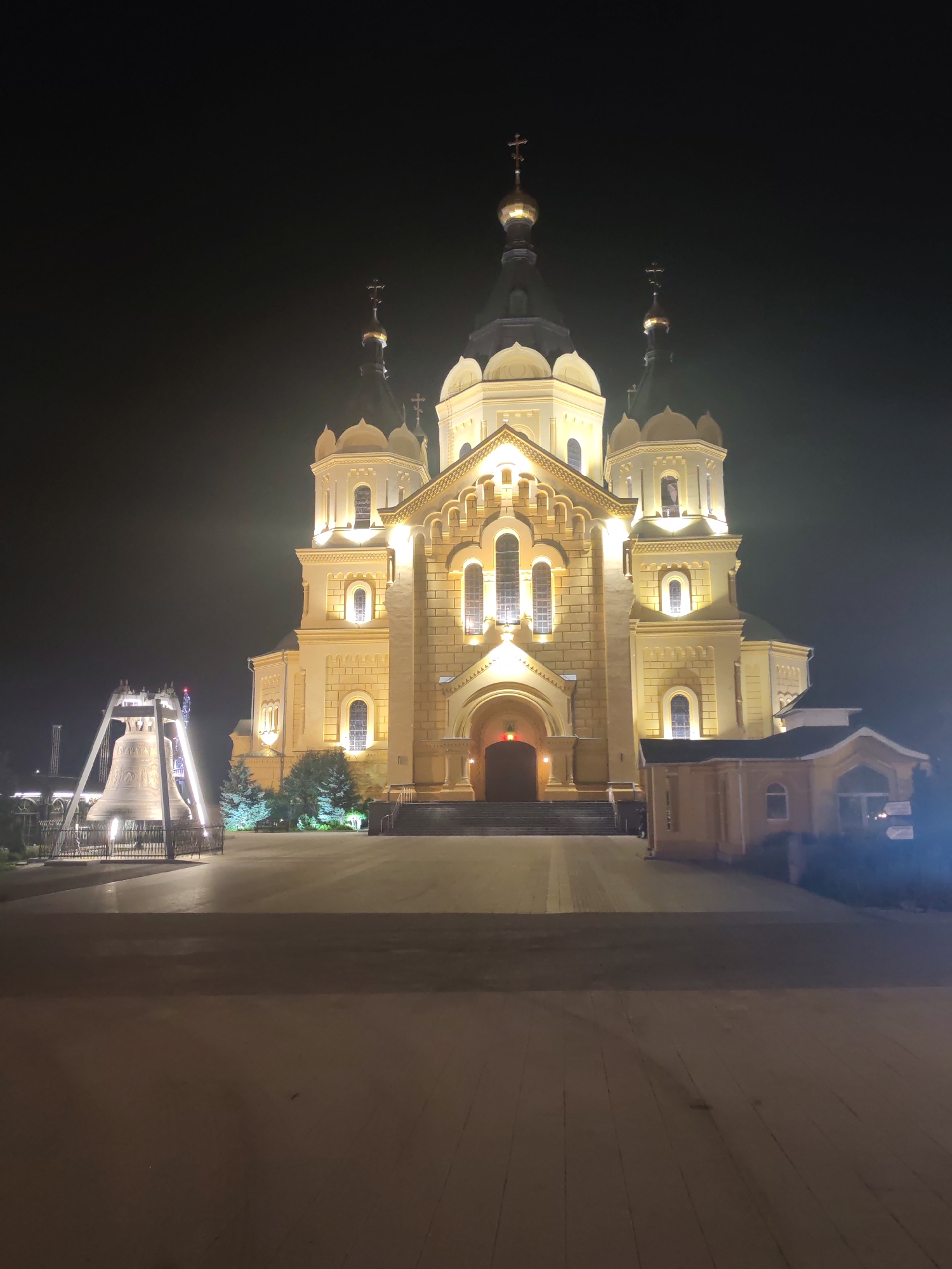 The image shows a large, illuminated church at night. The church is made of yellow stone and has multiple domes topped with crosses. It is lit from the outside, creating a warm glow. In the foreground, to the left, is a large bell mounted on a metal structure. The area in front of the church appears to be a paved courtyard. There are trees and some smaller buildings visible on the sides. The sky is dark, indicating nighttime. There are no people or visible actions in the scene. The scene is located in the city of Nizhny Novgorod, Russia.