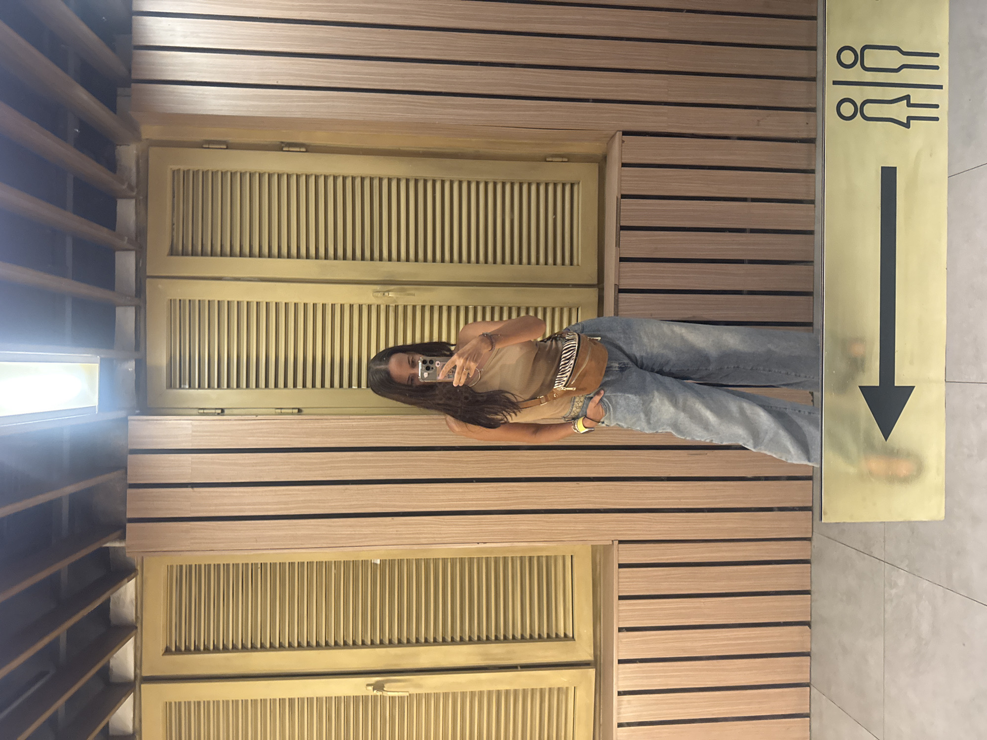 A woman is captured in a mirror selfie within an indoor setting. She holds a smartphone, partially obscuring her face, and wears a light brown tank top, light blue jeans, and a fanny pack with a striped strap around her waist. Her left wrist is adorned with a watch and bracelets. The background features a wall clad in horizontal wooden slats with dark dividing lines. Prominently in the center are golden-colored louvered panels, possibly doors or decorative screens. To the right, a vertical golden sign displays black pictograms indicating male and female restrooms at the top, followed by a large black arrow pointing downwards. The floor is covered with light-colored tiles. The scene is illuminated by artificial lighting, suggesting a commercial or public indoor environment. The presence of the restroom sign strongly indicates the location is within a public facility, possibly leading to or marking a restroom area in Kuta Selatan, Indonesia.