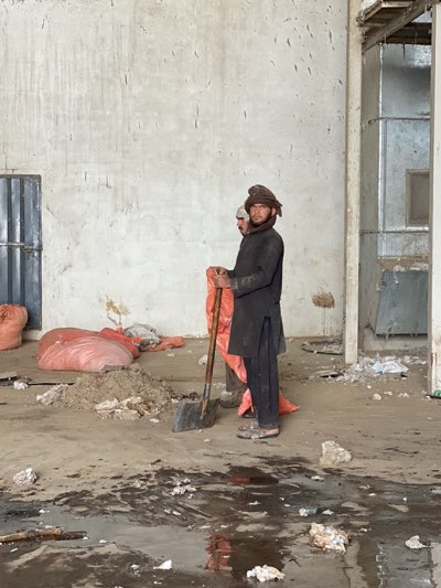 The image depicts two men standing in a dilapidated interior space. The man in the foreground wears a dark outfit, a head covering, and holds a shovel, while the man in the background wears similar clothing, his face partially obscured. The floor is littered with debris and appears damp. In the background, there are large bags and a pile of material. The walls are plain and aged, with evidence of disrepair. A metal structure is visible in the right background. The scene is consistent with the conditions often found in areas impacted by conflict or neglect. The image has been taken in a large hall and the environment suggests it is located in Kandahar, Afghanistan.