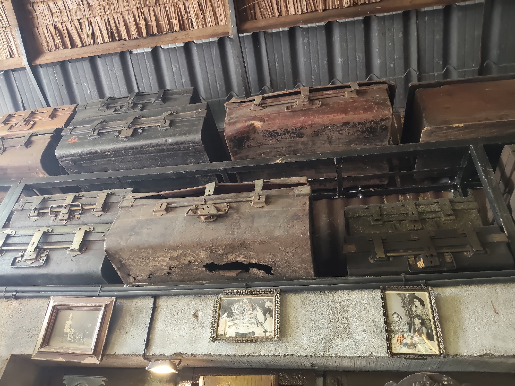 This is a low-angle, medium shot of several old, large metal trunks stacked on shelves. The trunks are various shades of gray and brown, and some appear to be rusty and damaged, with holes and dents. They have metal latches and handles. Above the trunks is a thatched roof. Below the trunks, on a concrete ledge, are three framed photographs. The photograph on the left is a sepia-toned image of a person on a bicycle. The middle photograph is a more detailed black and white image, and the photograph on the right is a black and white group portrait of four people. The scene appears to be indoors, possibly in a storage area or a historical exhibition space, with a corrugated metal ceiling visible. The lighting is dim, but the photographs are illuminated by what appears to be a spotlight. This image could be located in the Maimun Palace in Medan, Indonesia, a historical landmark known for its royal artifacts and architecture, which often include such antique items.
