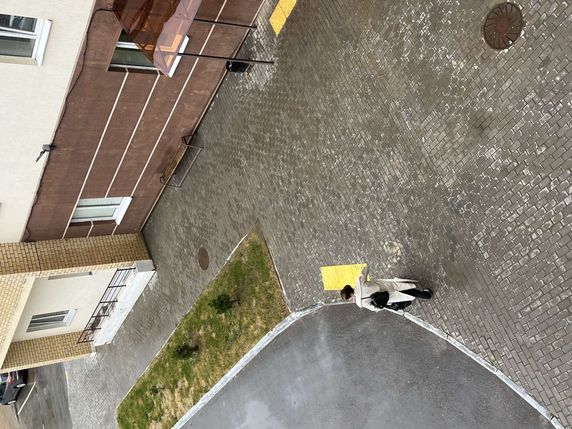 The image is an overhead shot taken in Chaikovsky, Russia. It shows a courtyard with brick paving, grass, and adjacent buildings. A woman in a light-colored coat stands on the pavement with a black bag. The pavement is wet, suggesting recent rain. There are manhole covers visible in the paved area and grass. The surrounding buildings are of various architectural styles, incorporating brick and plaster. A parked vehicle is visible in the corner of the image.