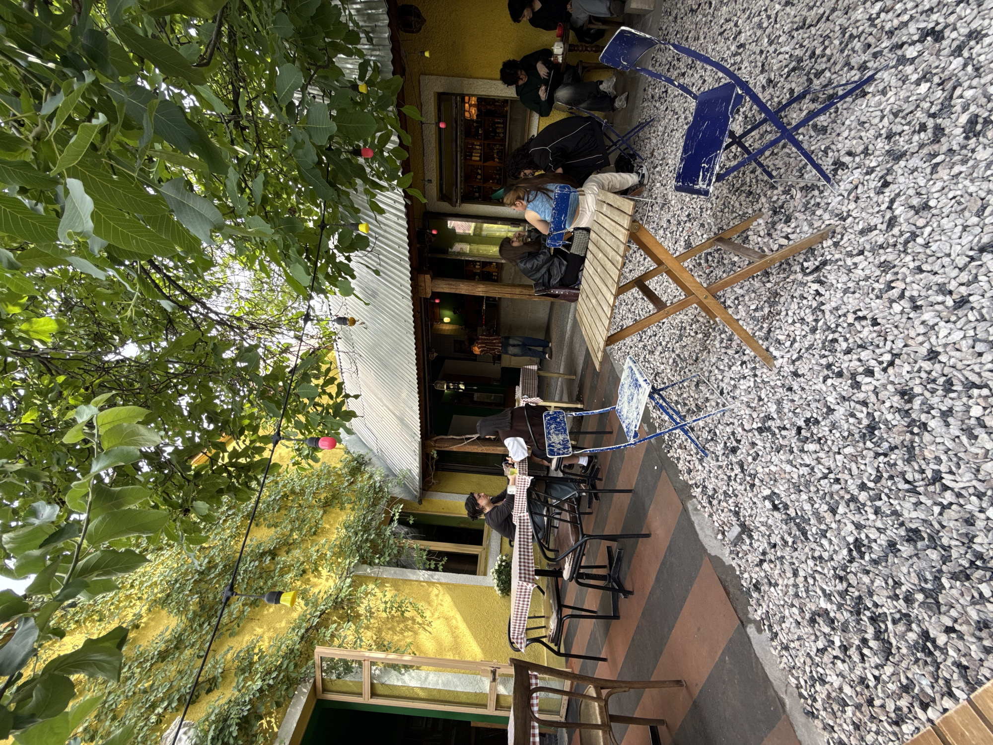 The scene is an outdoor dining area of a restaurant or cafe. Several people are sitting at tables, some working on laptops and others conversing. The location appears to be in Yerevan, Armenia, based on the architectural style. The dining area is partially covered by a corrugated metal roof. The ground surface varies, including gravel, striped tiles, and concrete. The building exterior is painted yellow and covered in climbing plants. Green foliage overhangs the scene, obscuring parts of the view. Decorative string lights are strung across the outdoor area. Tables are set with checkered tablecloths. The chairs are a mix of styles and colors.