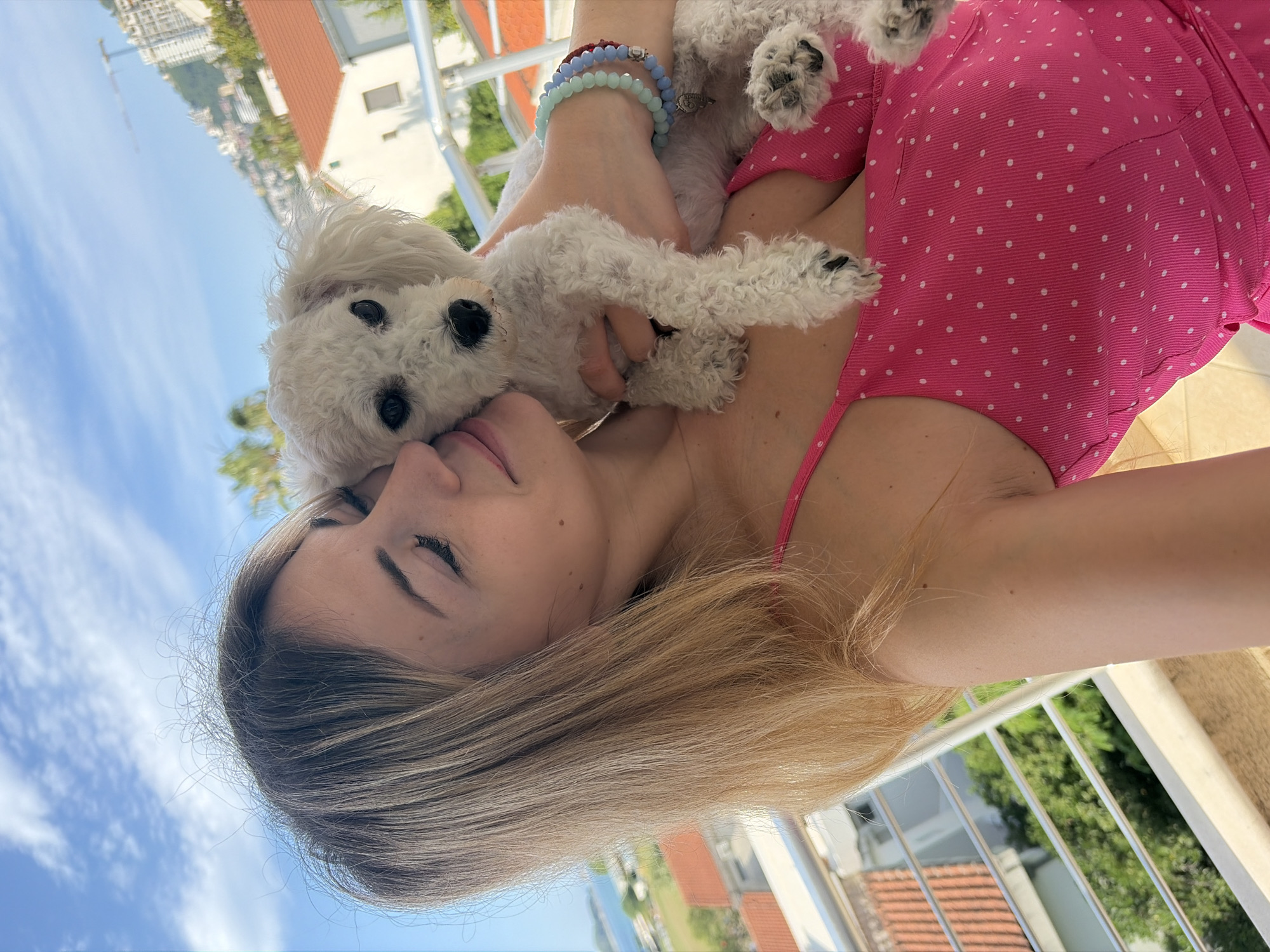 A woman with long blonde hair holds a small white dog. The woman wears a pink polka-dot dress. The dog is mostly white with black eyes and paws. A hand holding the dog wears bracelets. The background includes a balcony railing, trees, red rooftops, and the sky with scattered clouds. The location is Budva, Montenegro.