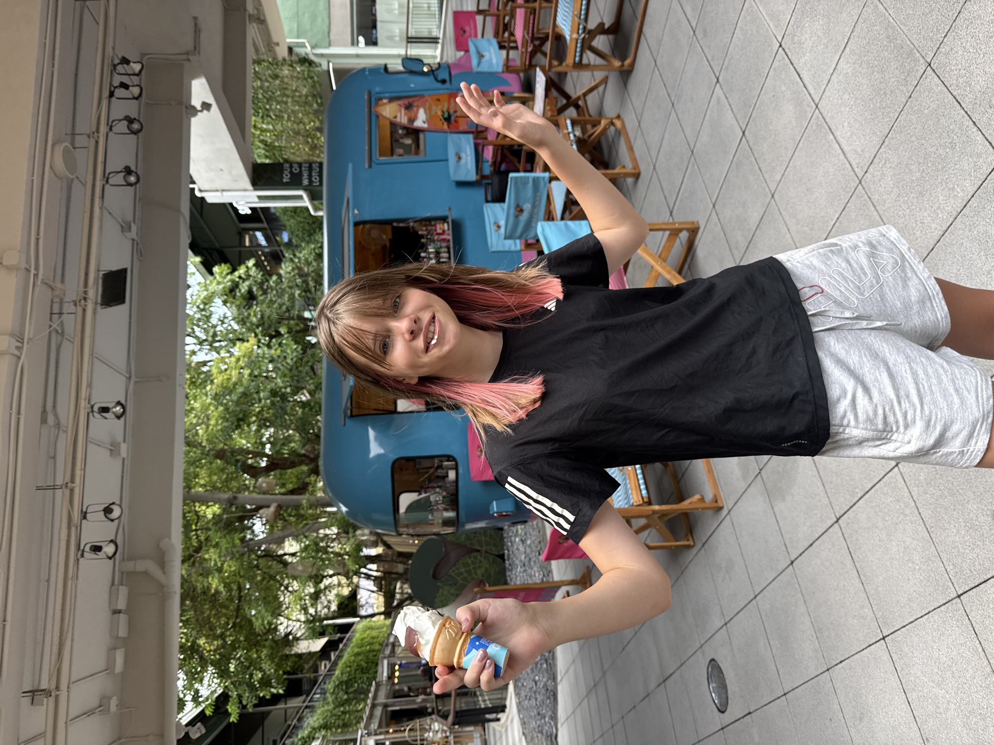 An undated, eye-level medium shot captures a young Caucasian female standing outdoors, holding an ice cream cone in her left hand. She has shoulder-length brown hair with pink-dyed tips and is smiling at the camera. She wears a black t-shirt with white stripes on the sleeves and light grey shorts. The background features a bright blue food truck with an open serving window, several colorful chairs, and lush green trees. A sign above the food truck reads "TOUR OF WHITE LOTUS". The ground is paved with square tiles. The setting appears to be an outdoor dining or recreational area in a tropical location, possibly a resort or a public space in Ko Samui District, Thailand.