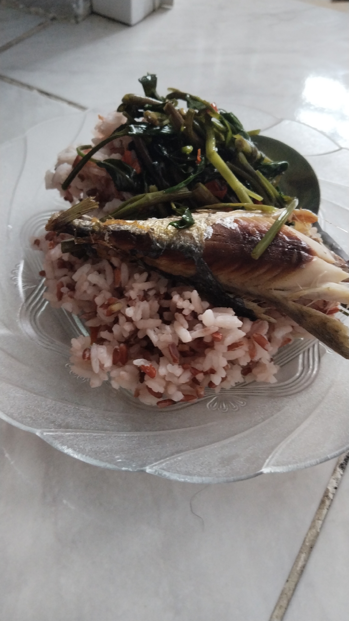 A plate of food containing rice, fish, and vegetables is displayed on a textured surface. The rice appears to be a mix of white and brown grains. A cooked fish, with its head and tail visible, rests on top of the rice. A portion of leafy green vegetables, likely stir-fried, is situated behind the fish and rice, partially on a dark green plate. The food is presented on a clear, ribbed glass plate. The background suggests an indoor setting with a tiled floor and a blurred wall. No people are visible in the image. The image is shot from a slightly elevated angle, focusing on the food.