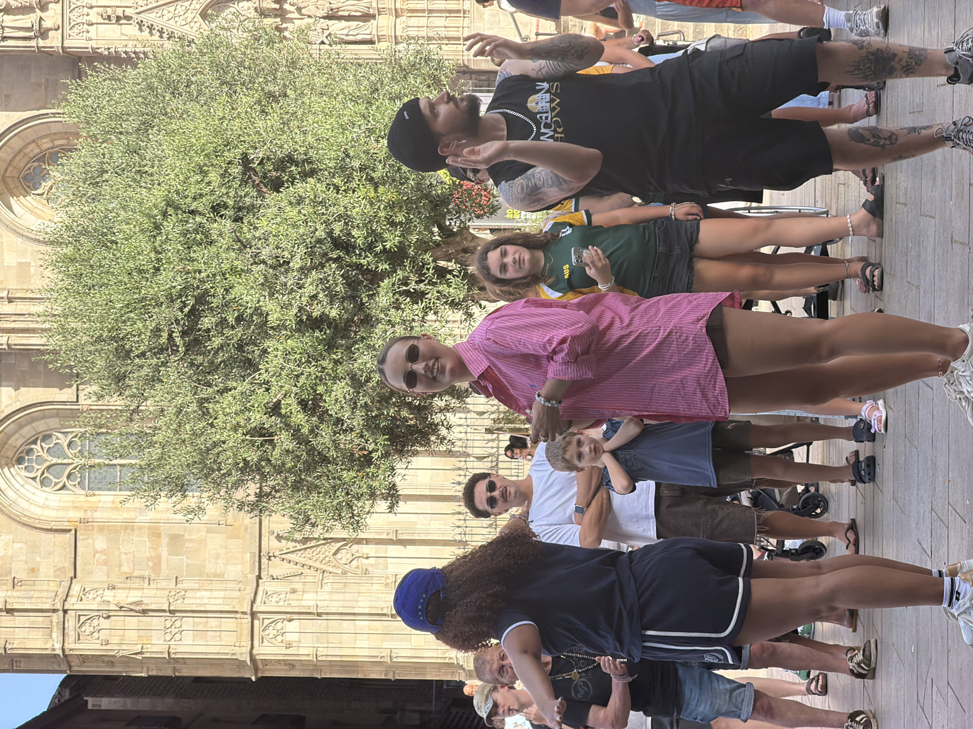 This is a medium shot of a group of people standing outside a large, ornate stone building with arched windows, likely a cathedral or a historic church, in Barcelona, Spain. The building's architecture suggests Gothic or Neo-Gothic style. The group is diverse in age and gender, with most individuals dressed in casual summer clothing.
In the foreground, a young woman in a pink striped shirt and dark shorts is looking forward, smiling, and holding a bracelet. To her left, a young boy in a blue shirt stands with his hands clasped, looking down. Behind him, a man in a white t-shirt and brown shorts stands with his arms crossed. Further left, a woman with curly dark hair wearing a blue cap and a dark t-shirt is gesturing with her arms, possibly addressing the group.
To the right of the young woman in the pink shirt, a young woman in a green and yellow sports jersey is holding a smartphone. Next to her, a man wearing a black beanie, a black sleeveless shirt with text, and dark shorts is gesturing with his hands. Other individuals are visible in the background, some partially obscured by the crowd and a large leafy tree. The ground is paved with stones. The overall atmosphere appears to be relaxed and public, possibly during a daytime event or gathering.