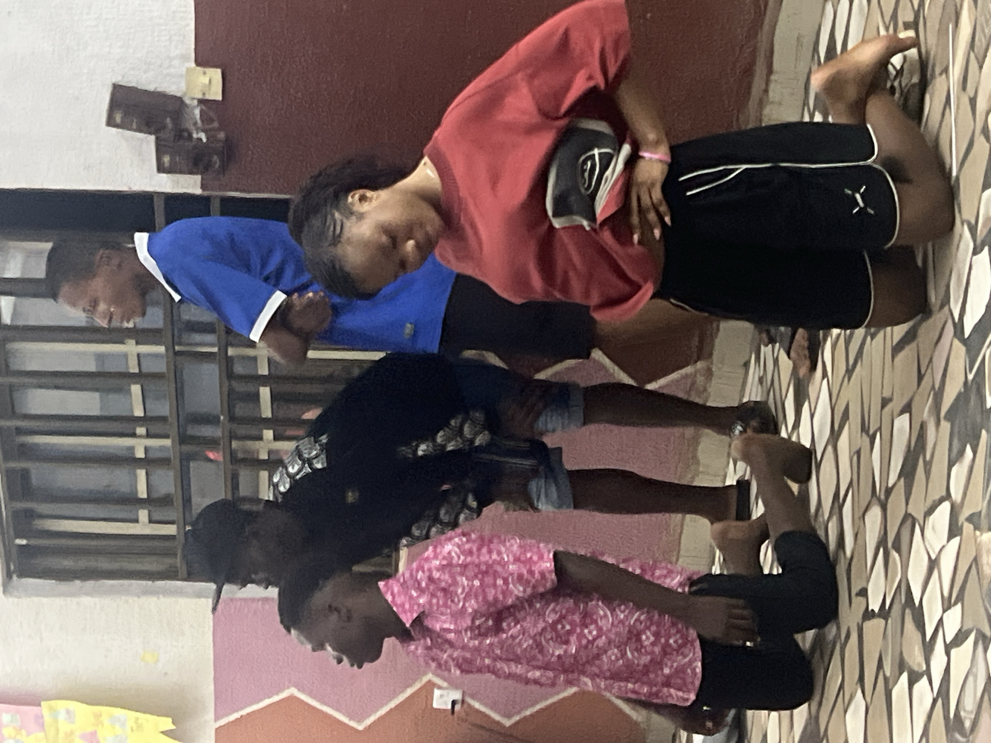 In Nkpor, Nigeria, four individuals are depicted indoors, kneeling on a patterned floor. A young woman in the foreground, wearing a red sweatshirt and black shorts, has her hand on her hip and is looking down. To her left, a young man in a pink patterned shirt also kneels, facing left. Behind him, another young man in a black t-shirt and denim shorts stands with his head bowed. Further back, a man in a blue shirt stands with his arms crossed, facing away from the camera. The room features a window with vertical bars, and a red wall with mounted speakers and light switches.