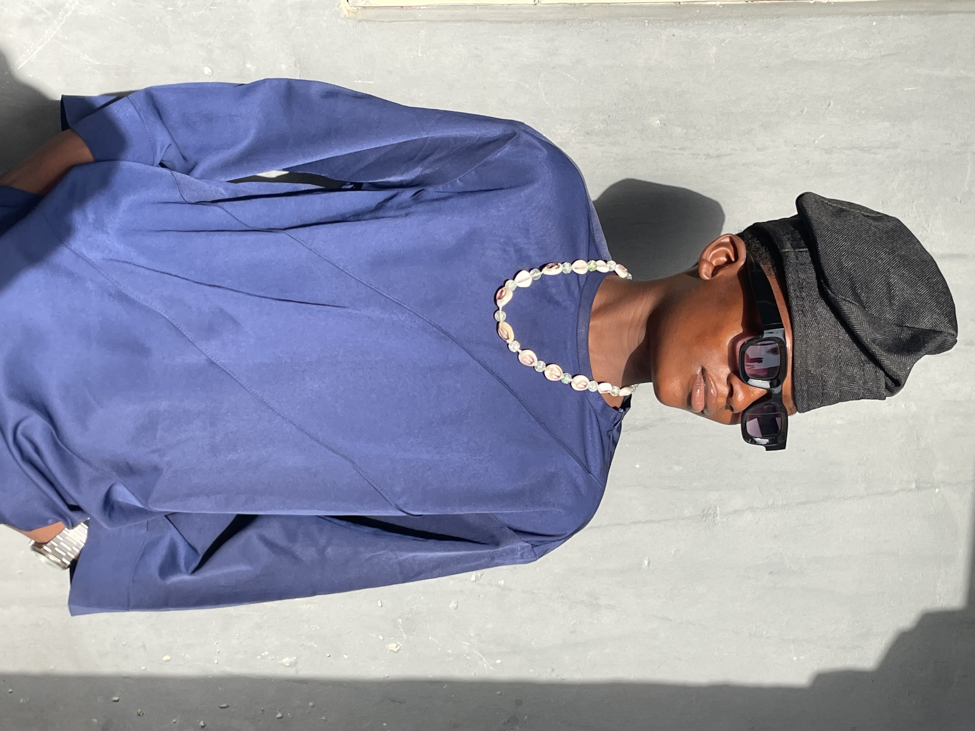 This image, taken in Badagry, Nigeria, features a person with dark skin posing outdoors in bright daylight. The individual is positioned against a light gray, textured concrete or paved surface, with a faint shadow cast behind them indicating strong sunlight.
The person is wearing a loose-fitting, long-sleeved top or tunic in a dusty blue or periwinkle color. They are accessorized with a dark gray or black fabric hat, possibly a beret or bucket hat style, worn on their head. Dark sunglasses with purple/pink reflective lenses obscure their eyes. Around their neck is a distinctive necklace composed of light-colored, shell-like beads interspersed with smaller, clear or light green spherical beads. A silver-toned watch is visible on their left wrist.
The person's head is tilted slightly downwards and to the side, suggesting a styled pose for the photograph. The lighting is direct and clear, characteristic of a sunny day. There is no discernible activity other than posing, and no visible text is present in the image. The background is simple and unadorned, focusing attention on the subject and their attire.